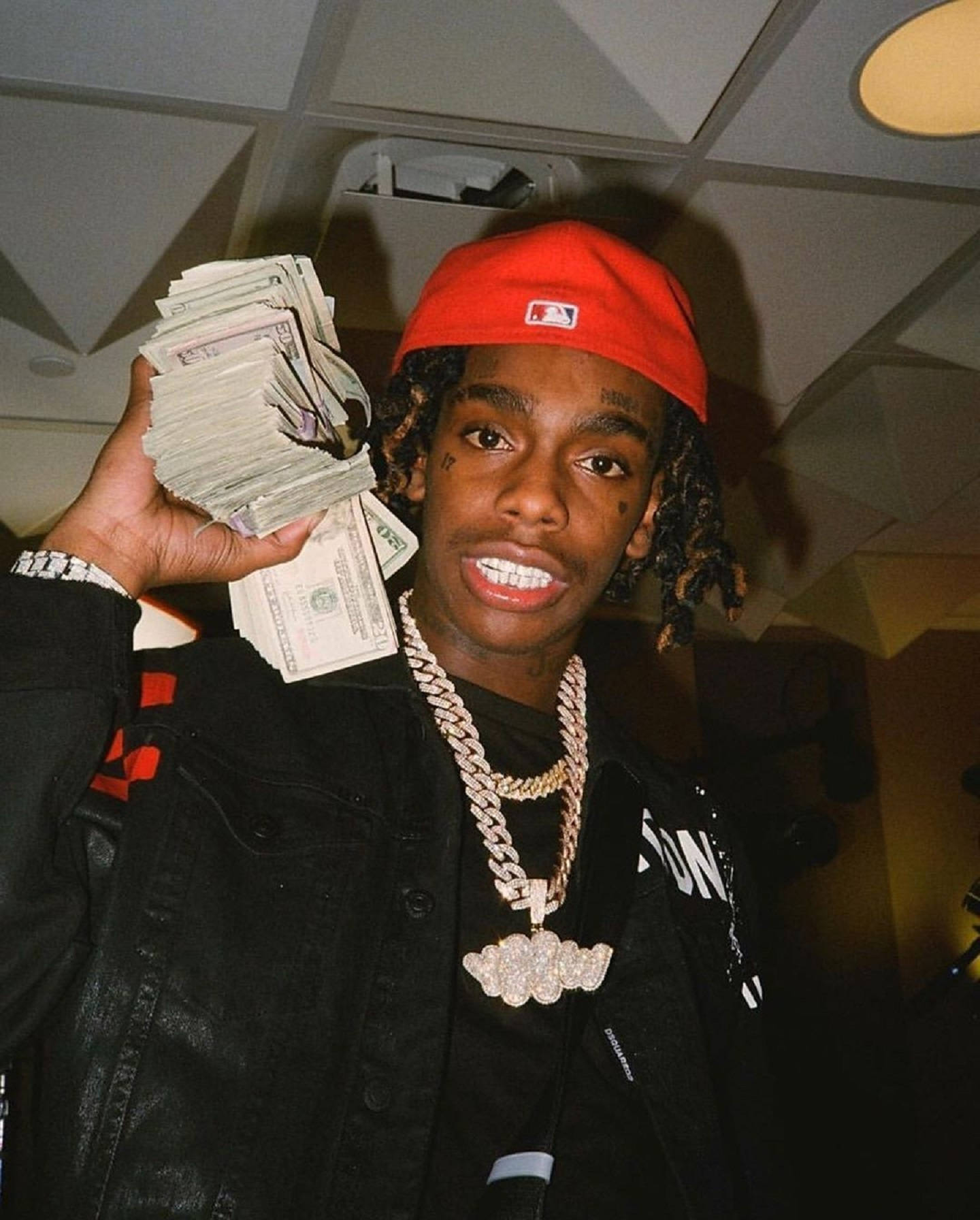 YNW Melly 嘻哈風格 壁紙 — 一名戴紅色帽、穿黑色外套的年輕男子自豪地展示一大疊現金，同時戴著一條粗鑽石鏈。