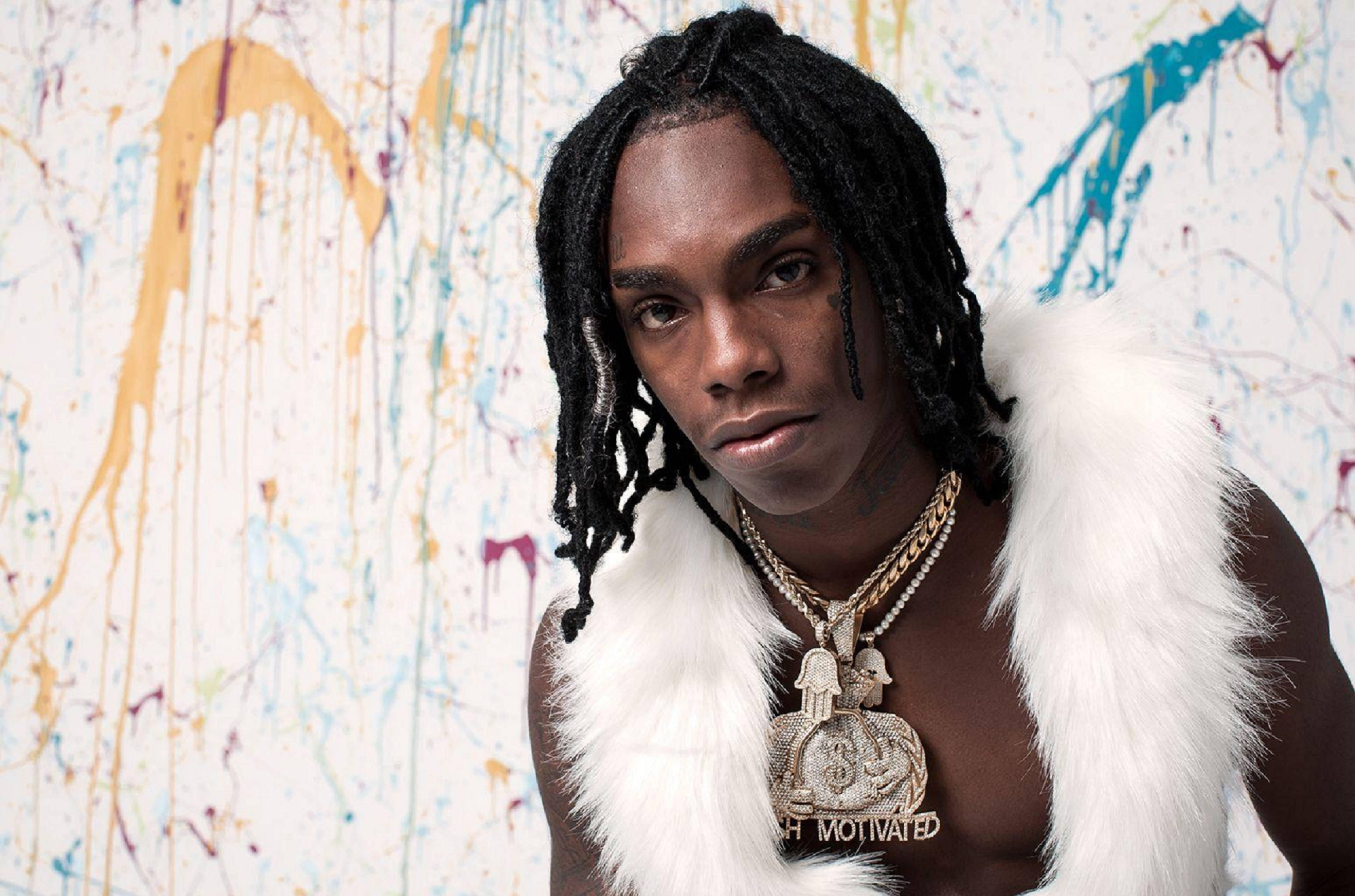 YNW Melly 榜單封面 壁紙 — 一個有辮子頭和戴金鏈的男人，穿著白色毛背心，站在色彩繽紛的油漆濺牆前。