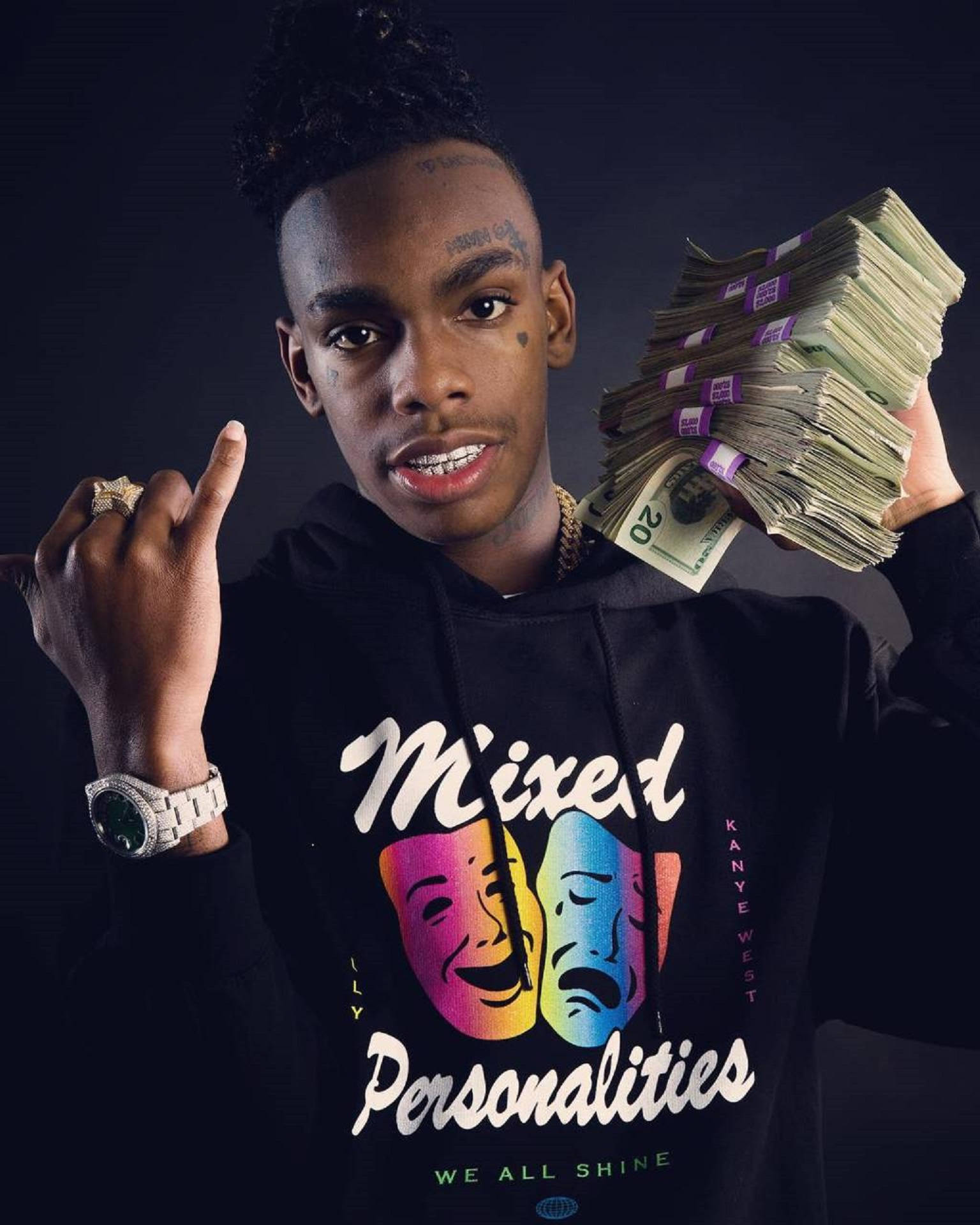 YNW Melly 混合主題 壁紙 — 一名身穿黑色連帽衫的年輕男子在黑暗背景前手持一疊疊現金並做出手勢。