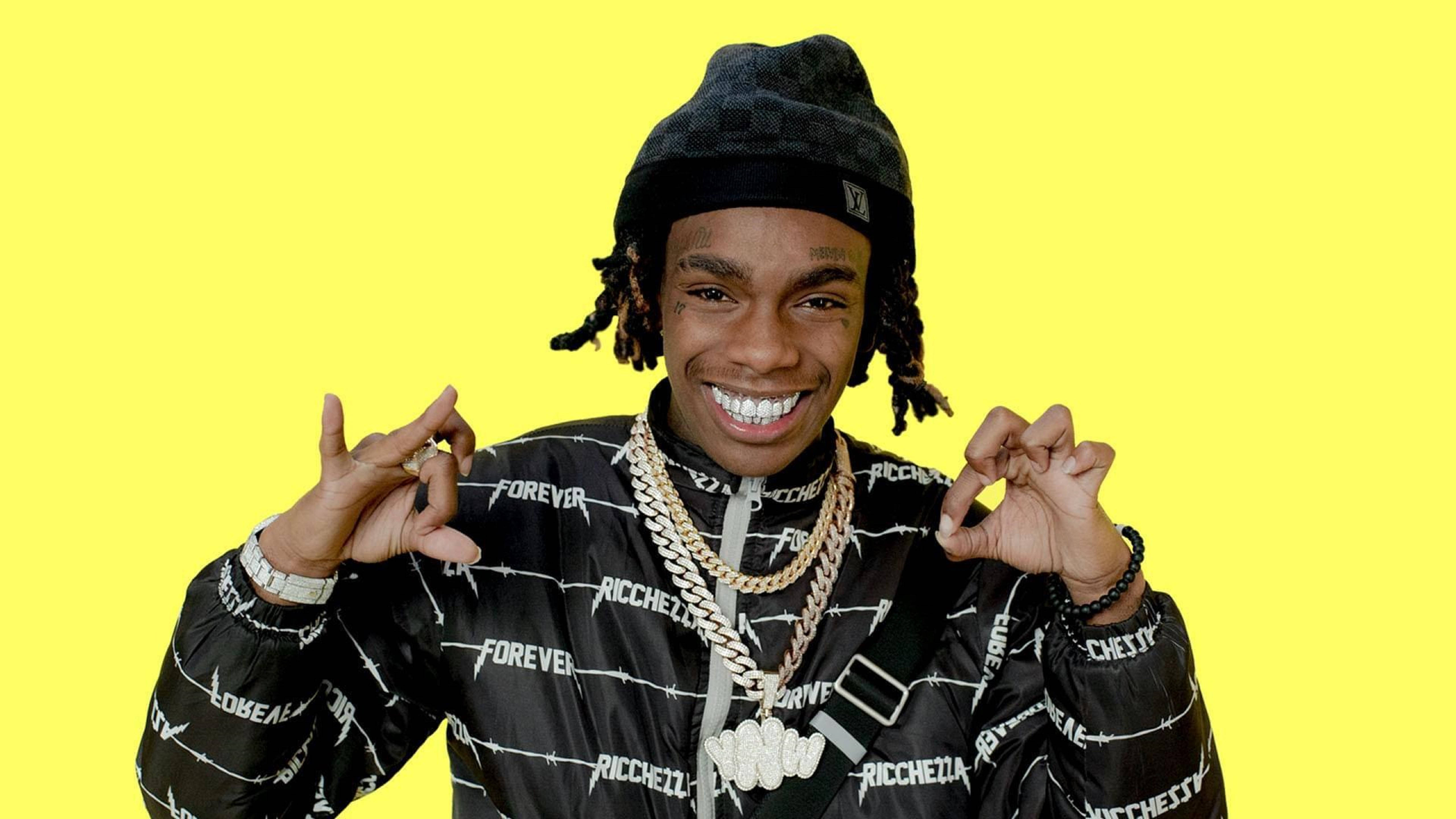 YNW Melly 混合人格 壁紙 — 一位微笑的年輕男子，梳著辮子並戴著冷帽，在明亮的黃色背景前自信地擺姿勢，身穿品牌外套和粗金鏈。