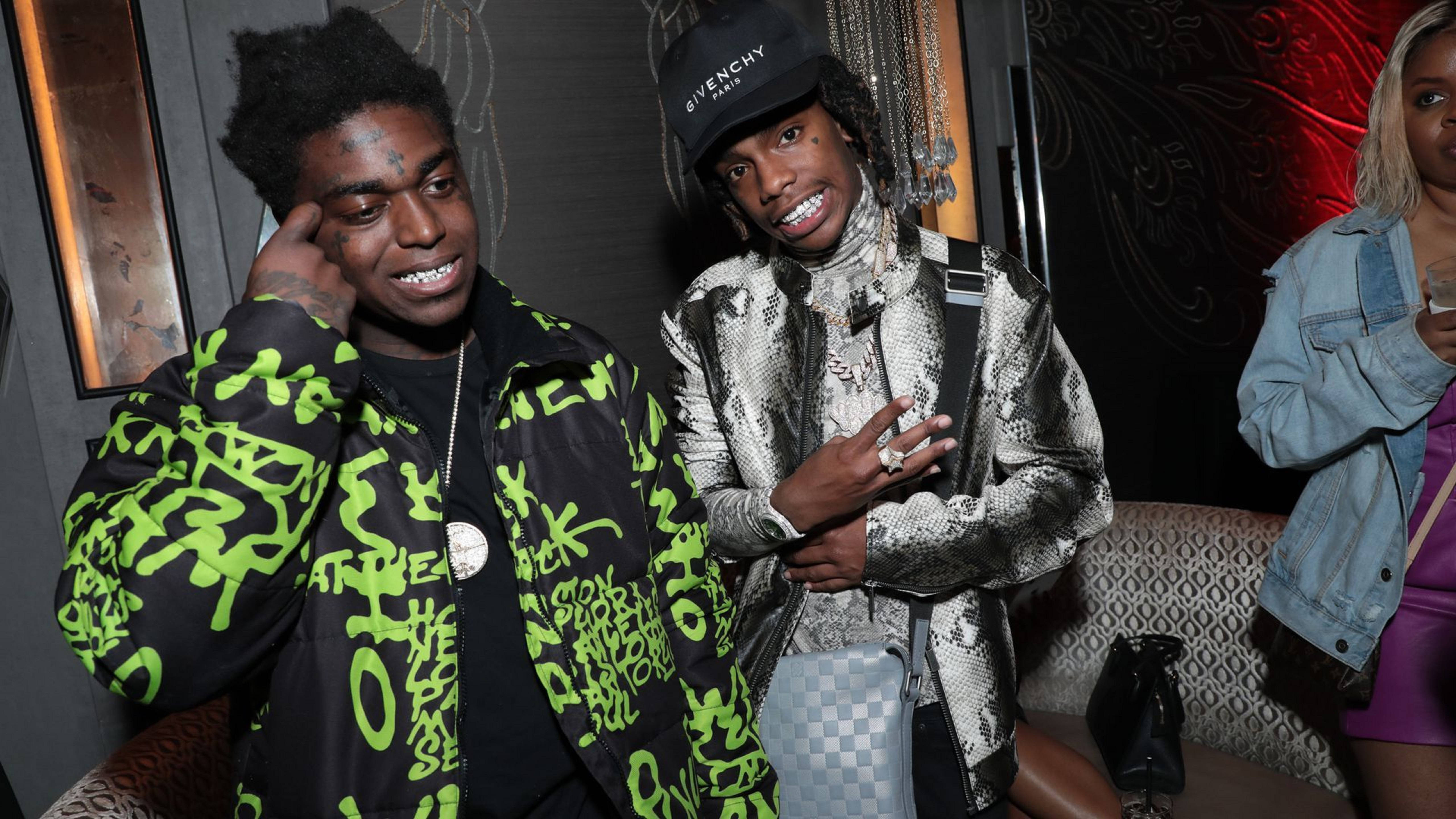 YNW Melly 與 Kodak Black 壁紙 — 兩位穿著時尚、身著大膽圖案外套的男士，在燈光昏暗、帶有紅色發光裝飾的俱樂部中自信地擺姿勢。