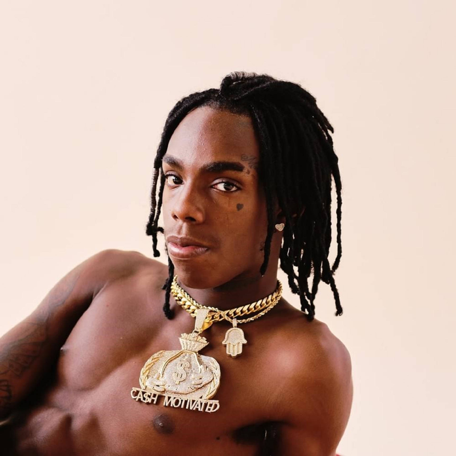 YNW Melly 與金項鍊的攝影棚寫真 壁紙 — 一個赤膊、梳著辮子的男人，戴著一條大型金色錢袋項鍊，上面寫著「CASH MOTIVATED」，背景是純米色。
