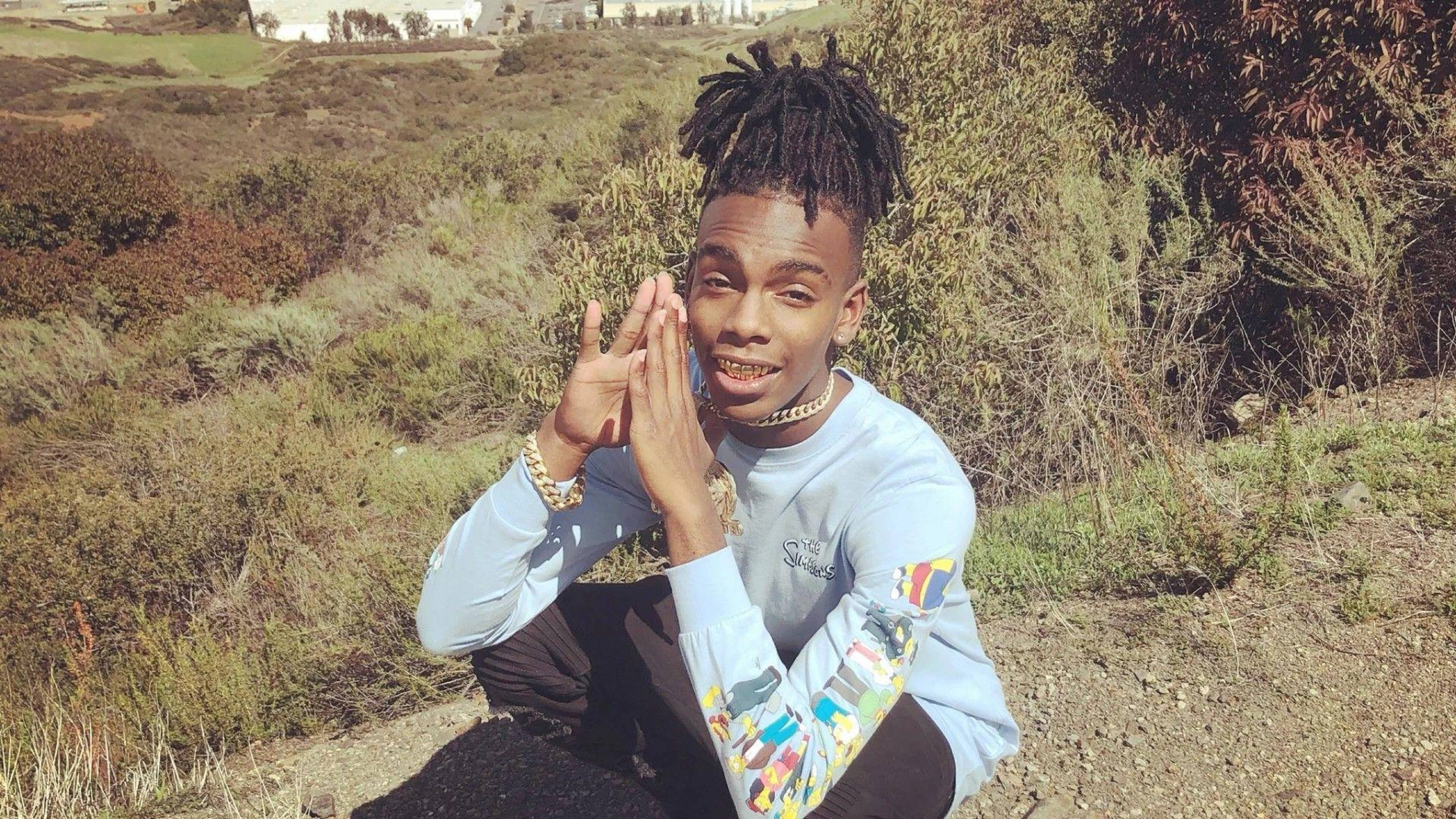 YNW Melly 草地復古風 壁紙 — 一位留著辮子的年輕男子蹲在戶外微笑，雙手靠近臉部。