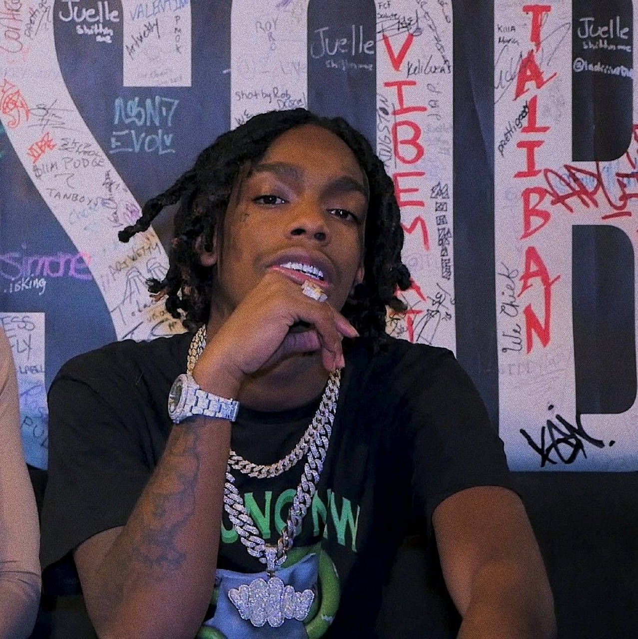 YNW Melly 街頭塗鴉牆肖像 壁紙 — 一位年輕男子編著辮子，佩戴鑽石首飾，在佈滿大膽紅白塗鴉標籤的牆前自信地擺姿勢。