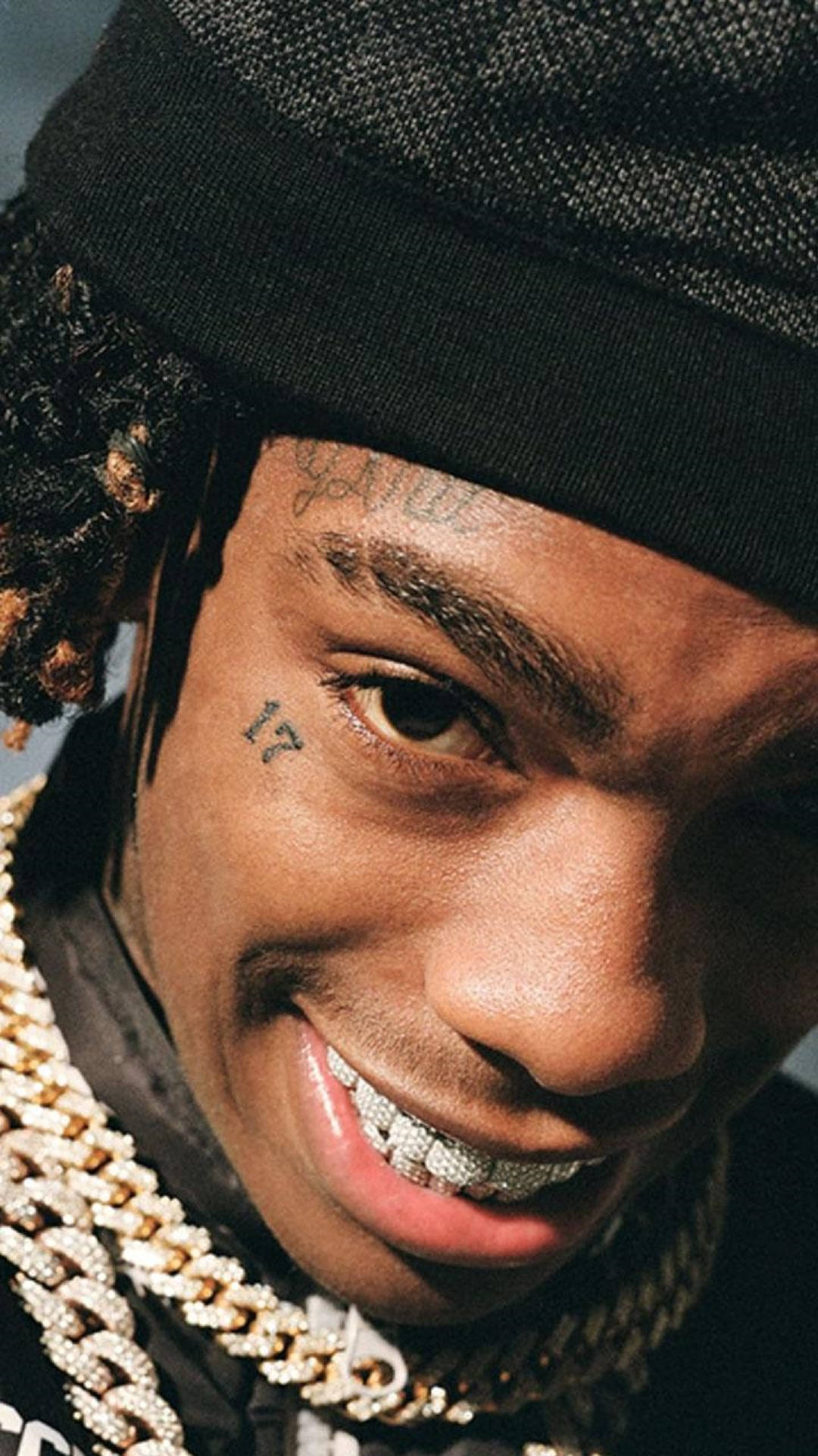 YNW Melly 近距離微笑特寫 壁紙 — 微笑男子的特寫肖像，帶有紋身和鑽石牙套，戴黑色無邊帽和粗金鏈。