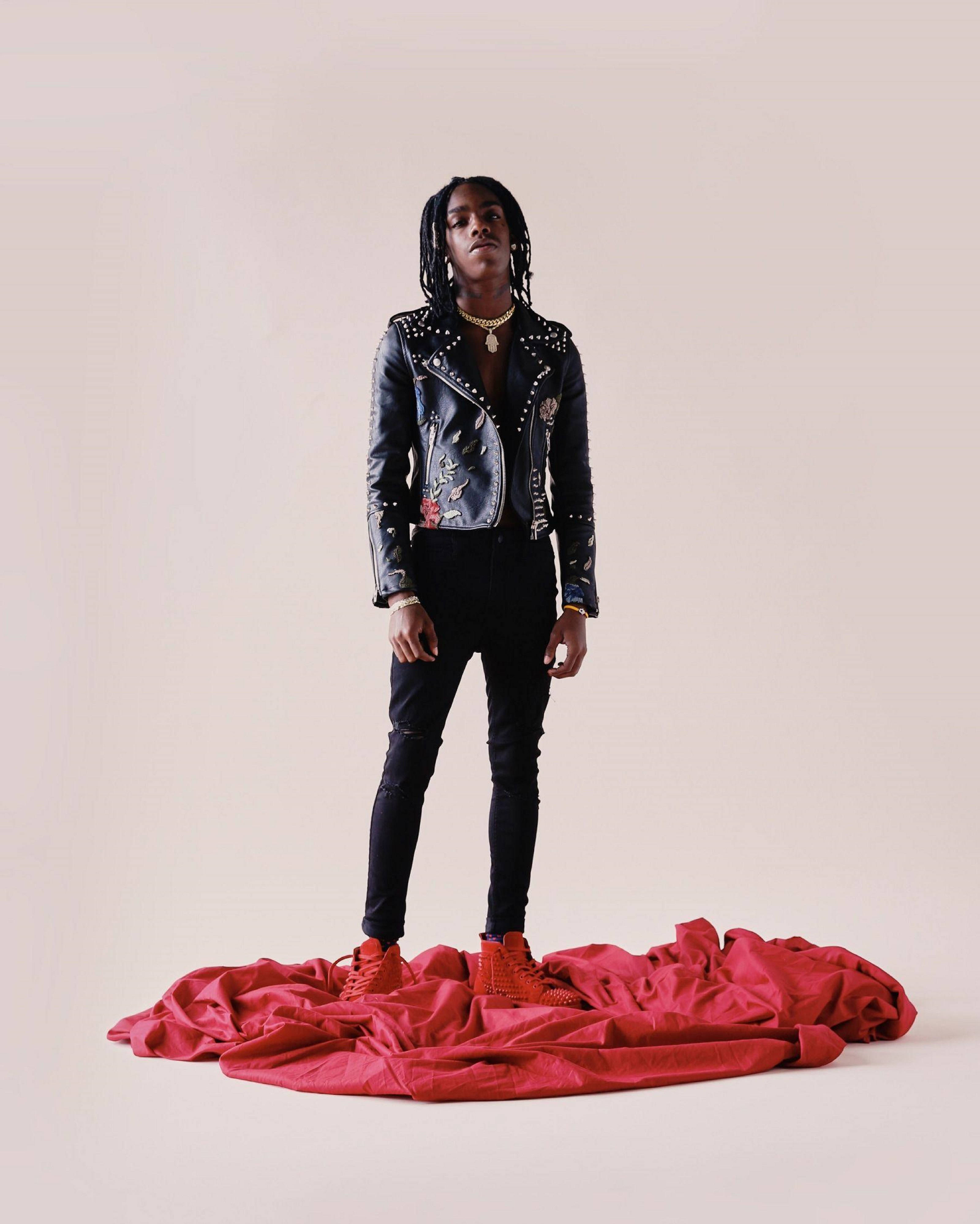 YNW Melly 革命外套攝影棚寫真 壁紙 — 一個留著辮子的人自信地站在一件飾有鉚釘的黑色皮褸和紅色運動鞋上，腳下是皺巴巴的紅色布料，背景是純色米色。
