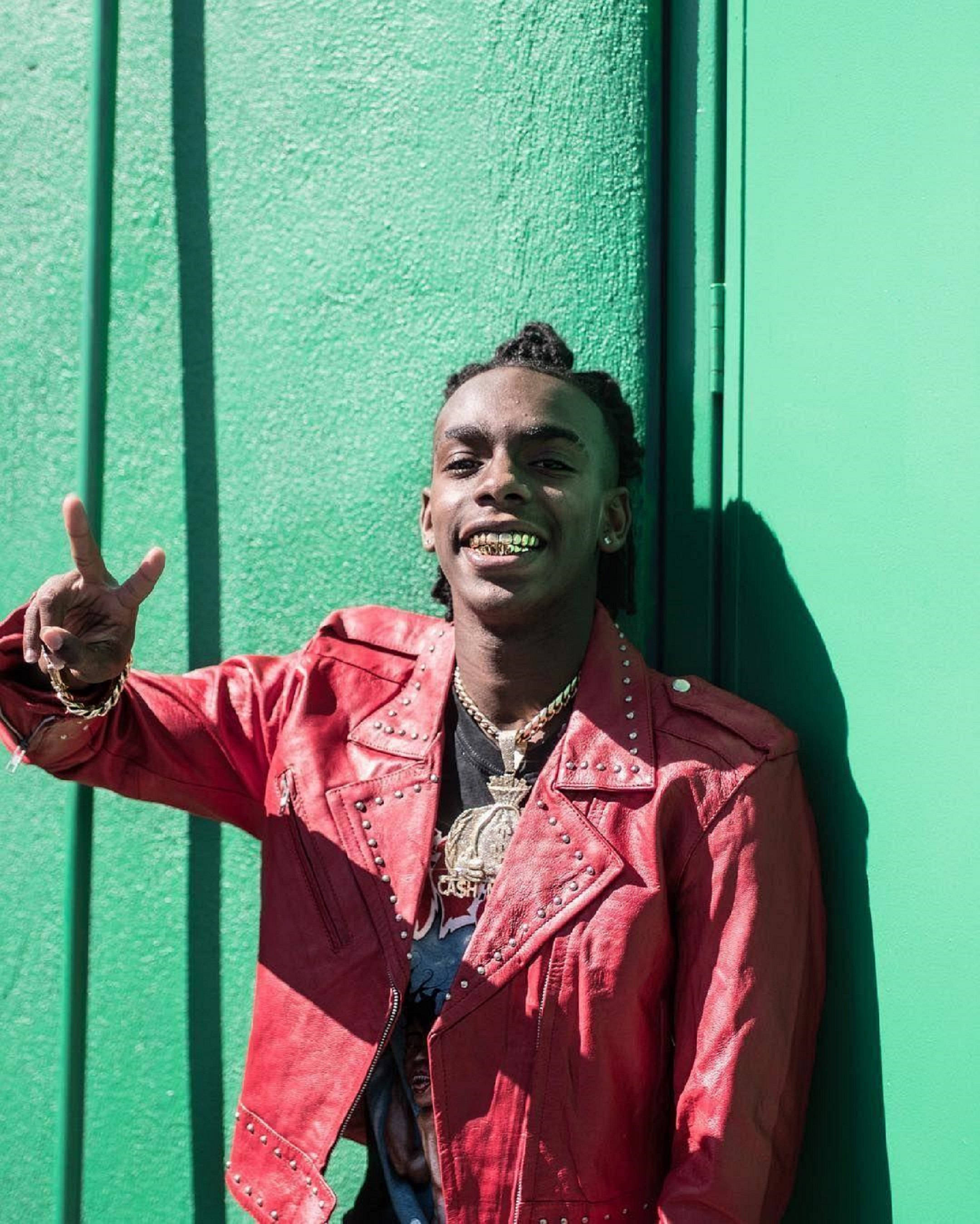 YNW Melly 魔鬼剋星造型 壁紙 — 一位身穿紅色鉚釘皮褸的年輕男子在明亮陽光下對著充滿活力的綠色牆壁微笑並做手勢。