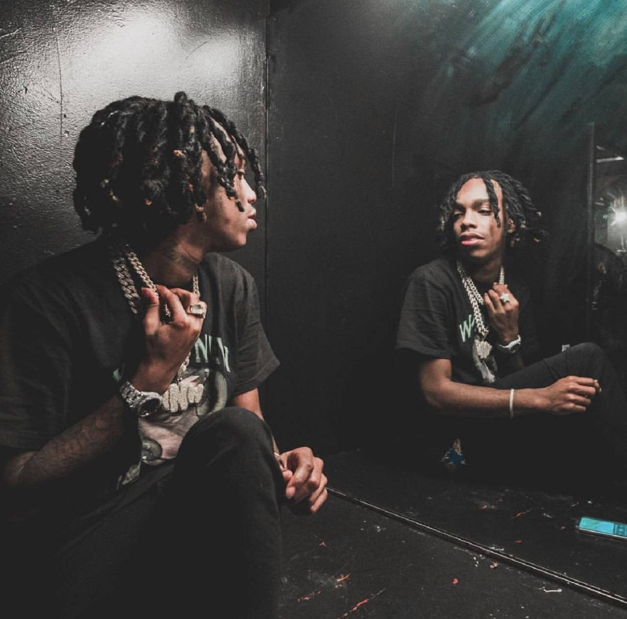 YNW Melly 黑色系 壁紙 — 一位年輕的雷鬼辮男子坐在燈光昏暗的房間裡，調整他的鏈條項鍊，同時凝視著鏡中的倒影。