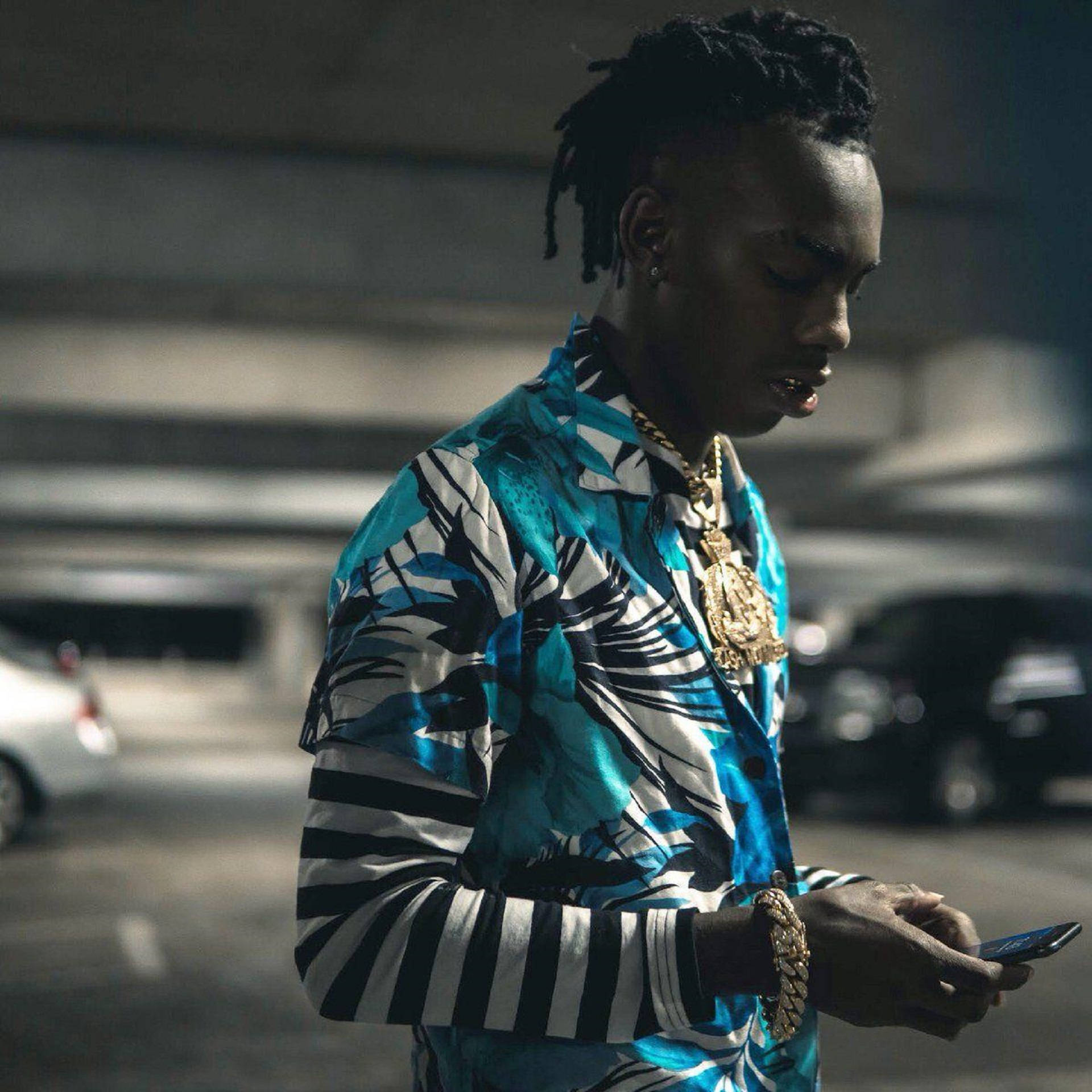 YNW Melly專輯預告 壁紙 — 一位時尚男士穿著大膽的藍黑圖案襯衫，在昏暗的停車場燈光下查看手機。