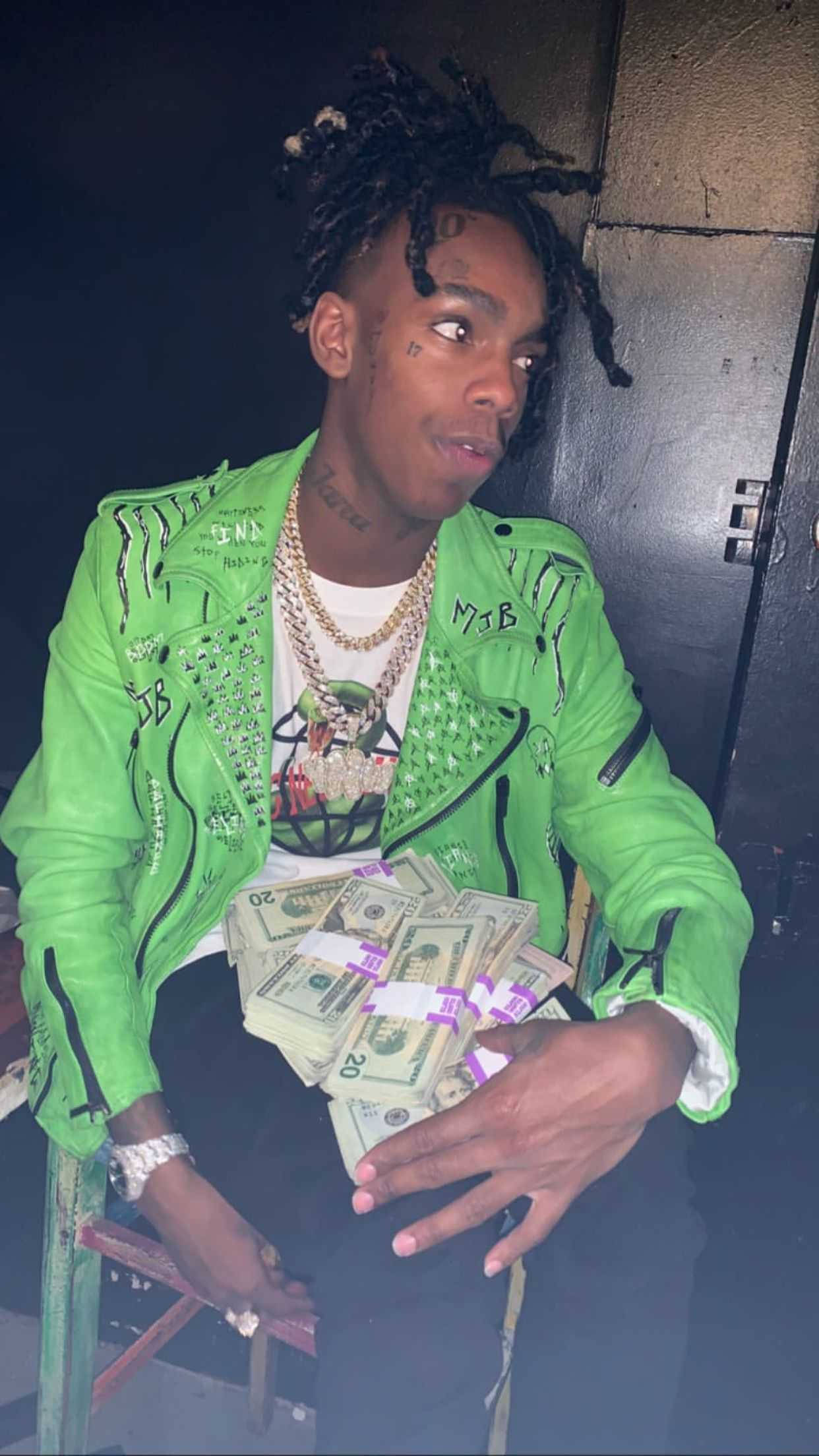 YNW Melly演唱會 壁紙 — 一位留著辮子的年輕男子身穿亮綠色外套，在昏暗的房間裡拿著一疊疊美國二十美元鈔票。
