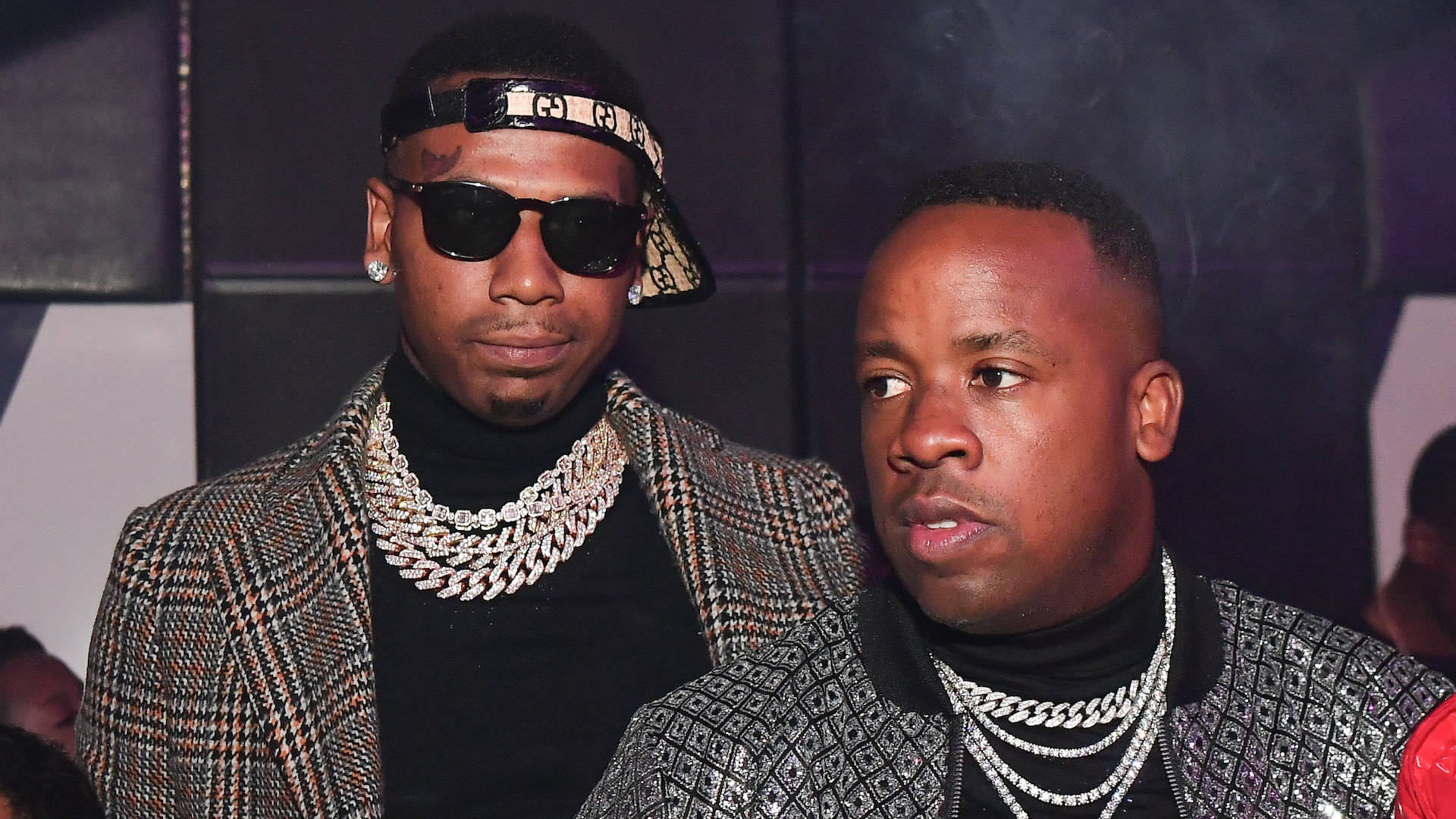 Yo Gotti 與 Moneybagg Yo 壁紙 — 兩位時尚男士身穿圖案外套和粗鏈條，在燈光昏暗、背景有煙霧的活動中自信地擺姿勢。