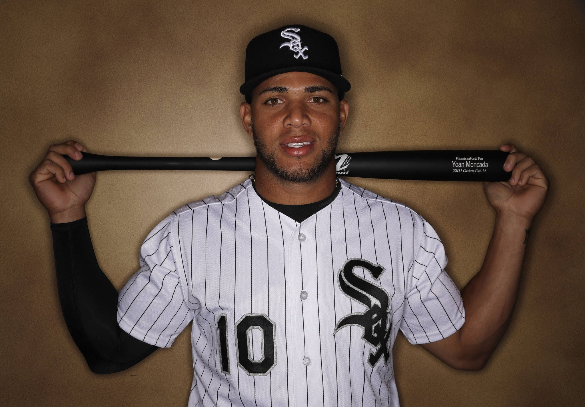 Yoan Moncada 手持棒球棒擺姿勢 壁紙 — 一名身穿芝加哥白襪隊球衣的棒球手，在純棕色背景前將球棒扛在肩上。