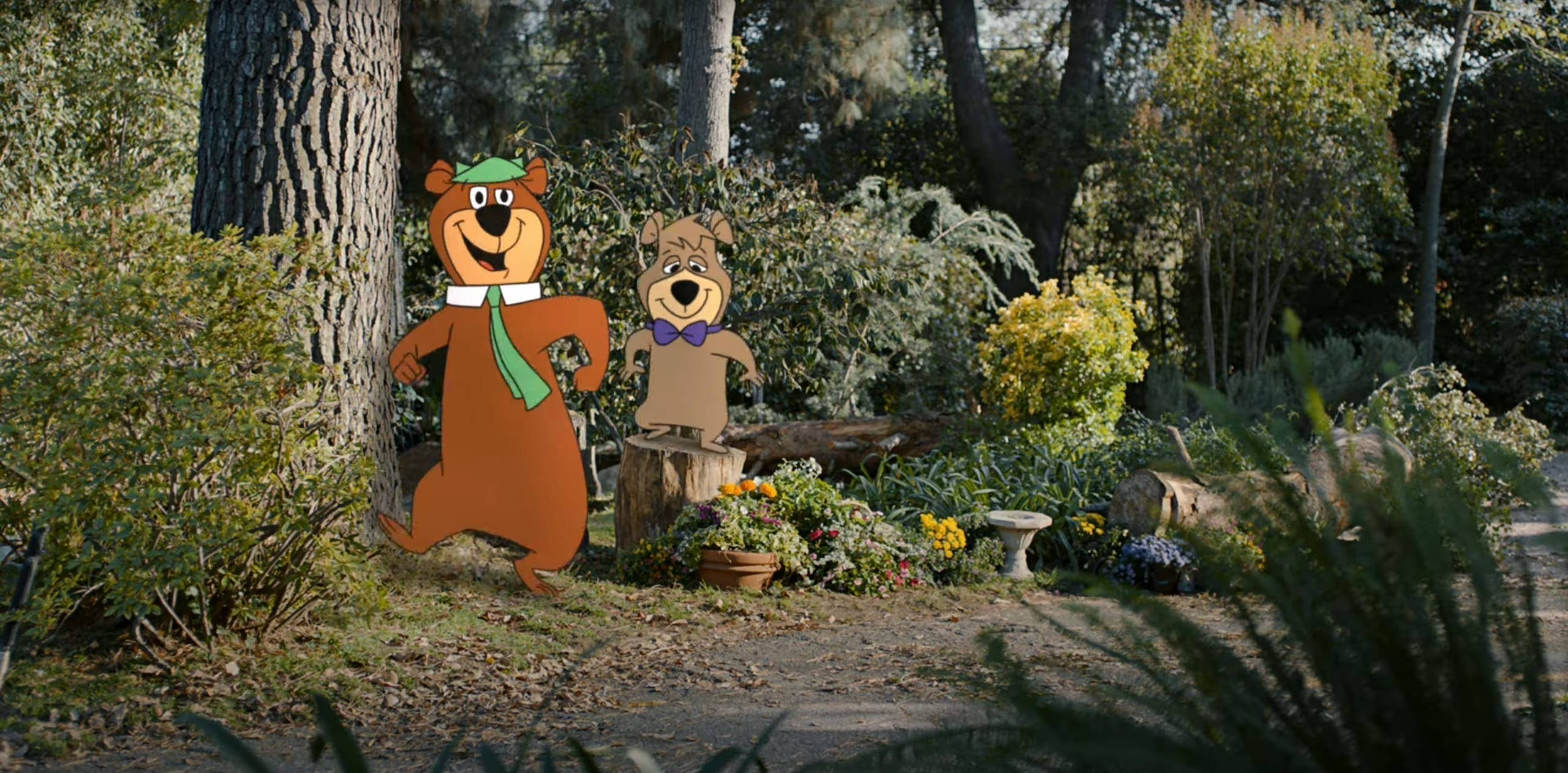 Yogi Bear GEICO廣告 壁紙 — 史酷比和沙吉的卡通剪影站在陽光明媚的花園中，有樹木、花朵和一個石鳥浴盆。