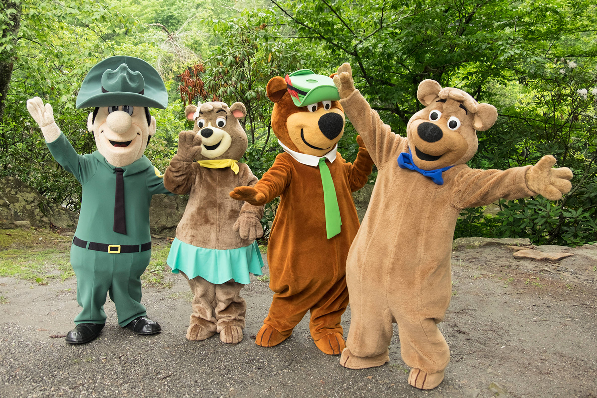 Yogi Bear 與朋友們 壁紙 — 四個穿著吉祥物服裝的開心卡通人物在綠樹成蔭的森林小徑上快樂地揮手。