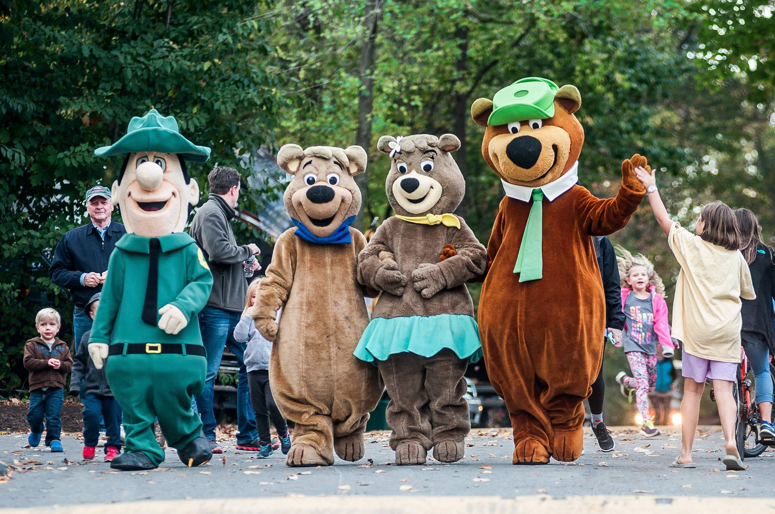 Yogi Bear與朋友吉祥物 壁紙 — 四個穿著服裝的快樂卡通熊角色在綠葉成蔭的公園裡與孩子們互動。