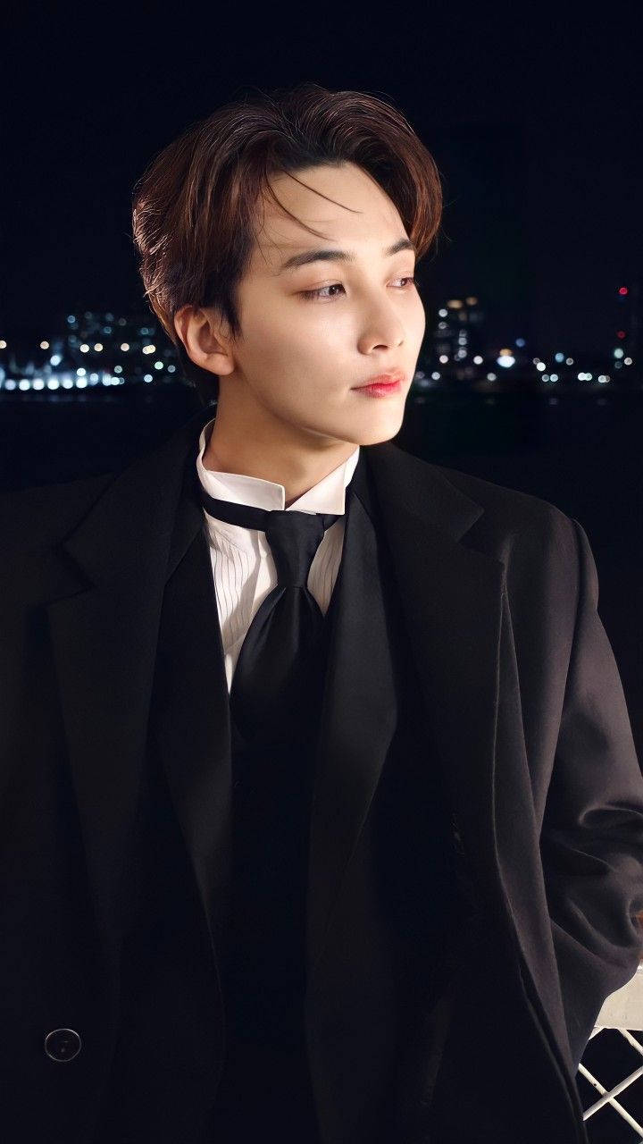 Yoon Jeonghan 西裝造型 壁紙 — 一位身穿黑色西裝的年輕男子，在模糊的城市夜景和閃爍燈光中若有所思地凝視。