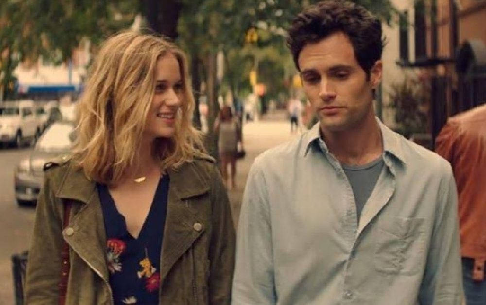 《You》Netflix劇集中的喬·高登與貝克 壁紙 — 一位微笑的女子穿著綠色外套，與一位穿著淺色襯衫、沉思的男子並肩走在綠樹成蔭的城市街道上。