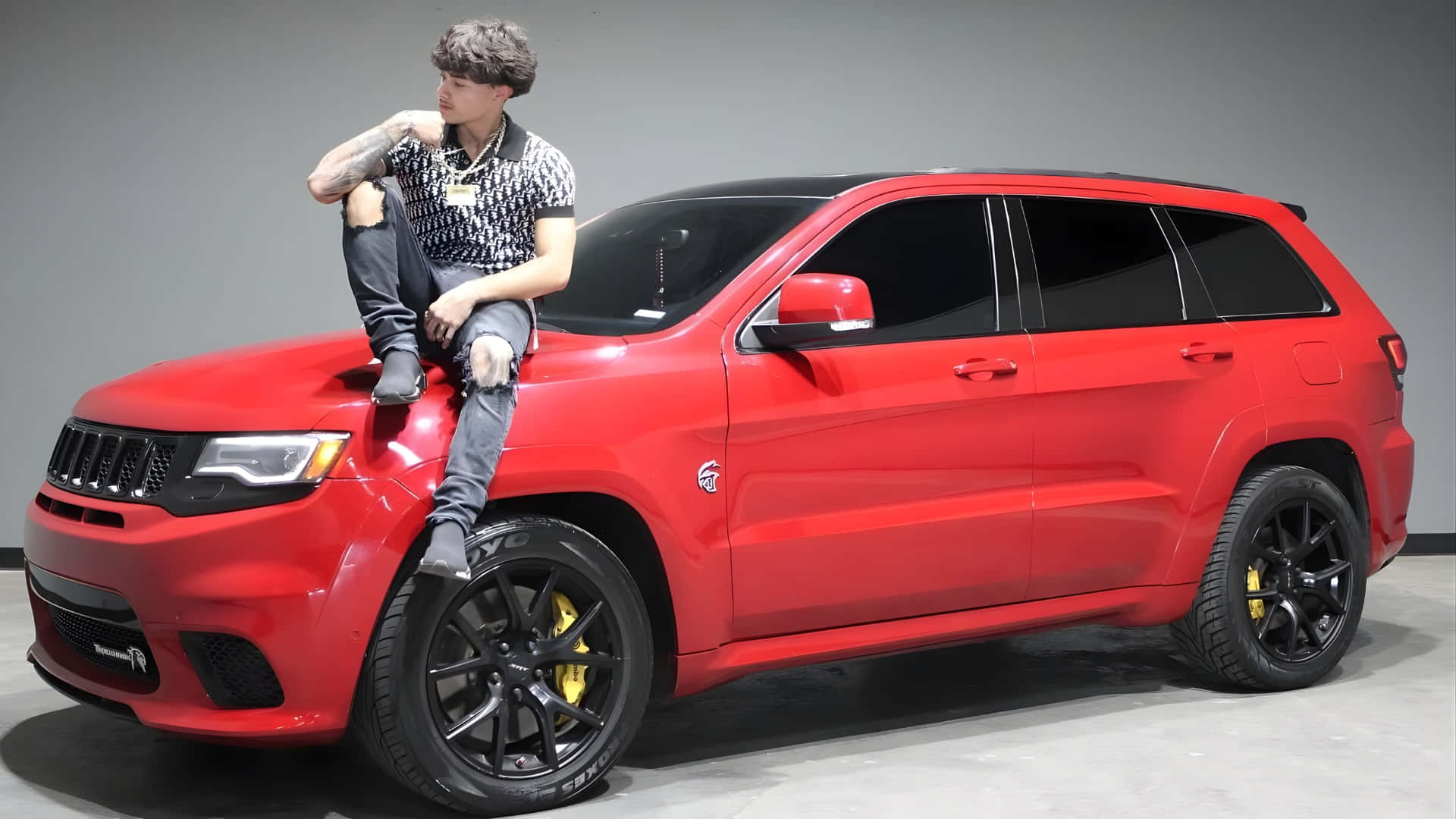  壁紙 — 一位有紋身的年輕男子隨意地坐在一輛鮮紅色Jeep Grand Cherokee上，車子配有黑色輪圈和黃色制動卡鉗，背景是純灰色攝影棚。