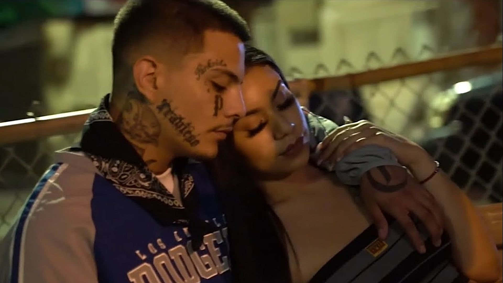  壁紙 — 一個有紋身的男人和女人在溫暖的夜燈下，在鐵絲網圍欄附近溫柔地擁抱。