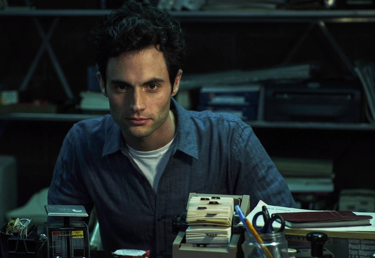 《You》劇集演員 Penn Badgley 壁紙 — 一個深色捲髮的男人坐在燈光昏暗的辦公室裡，面對雜亂的書桌，專注地凝視前方。