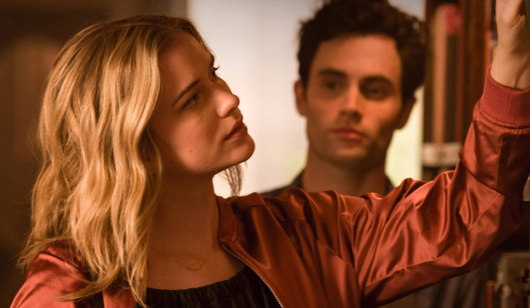《You》劇集演員 Penn 與 Ambyr 壁紙 — 一位身穿紅色緞面外套的女性向上伸手，而一名男性在溫暖燈光的室內場景中專注地注視著。