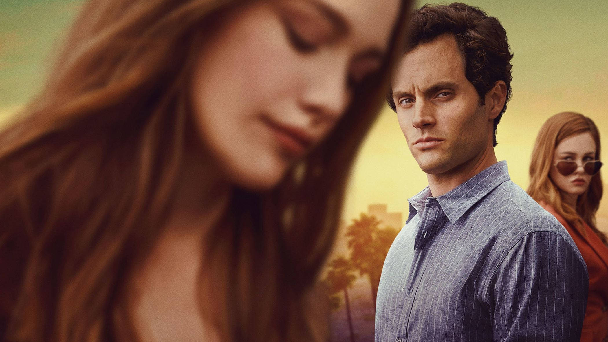 《You》劇集角色 Joe Goldberg 與 Love Quinn 的冷峻瞬間 壁紙 — 一名穿著條紋襯衫的男子站在兩個女人之間，一個在前景模糊，另一個戴著太陽眼鏡，沐浴在溫暖的金色夕陽光中。