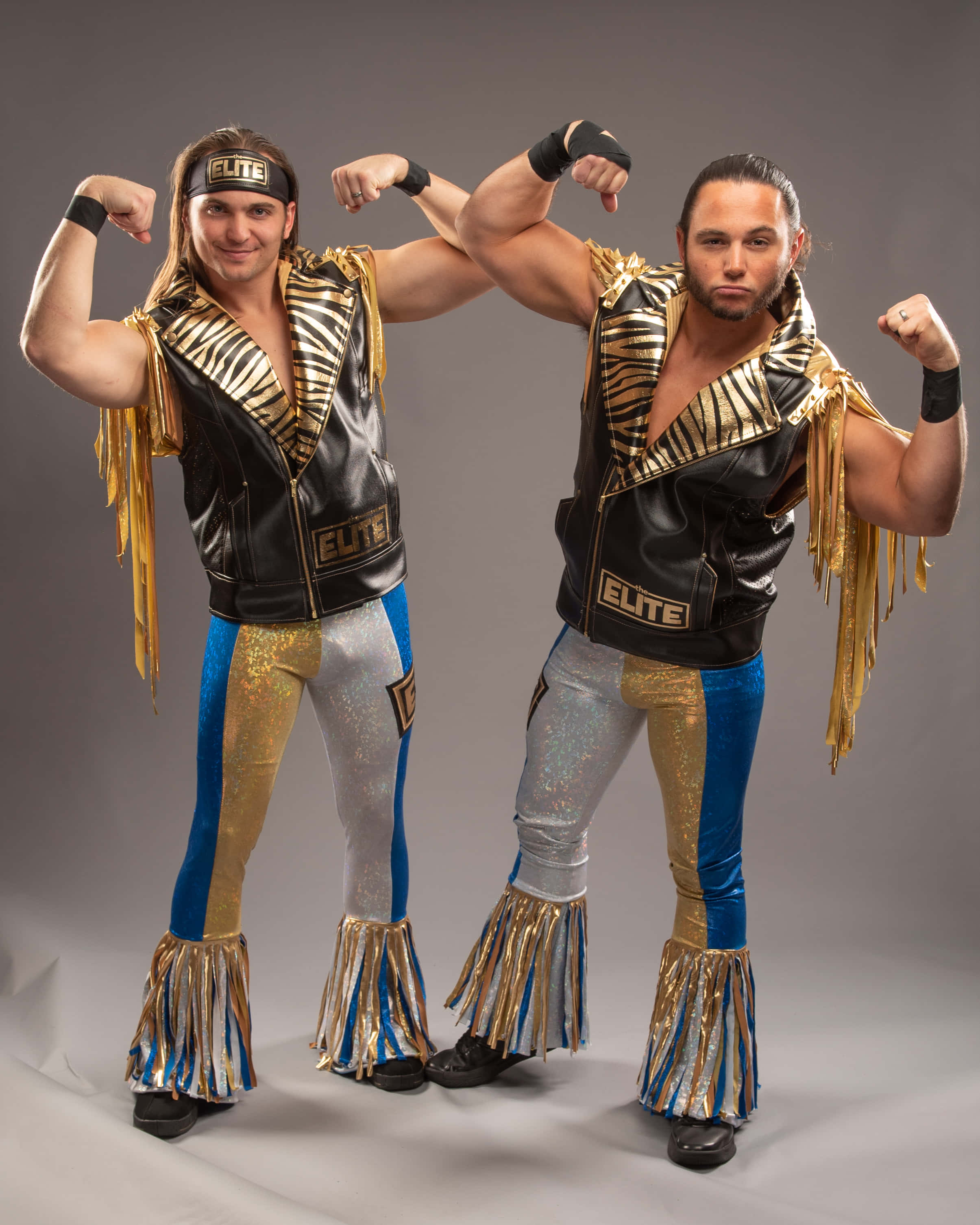 Young Bucks 尼克與馬特·傑克森拍攝照 壁紙 — 兩名肌肉發達的摔角手穿著閃亮的金色、藍色和黑色服裝，在樸素的灰色工作室背景前自信地擺姿勢。