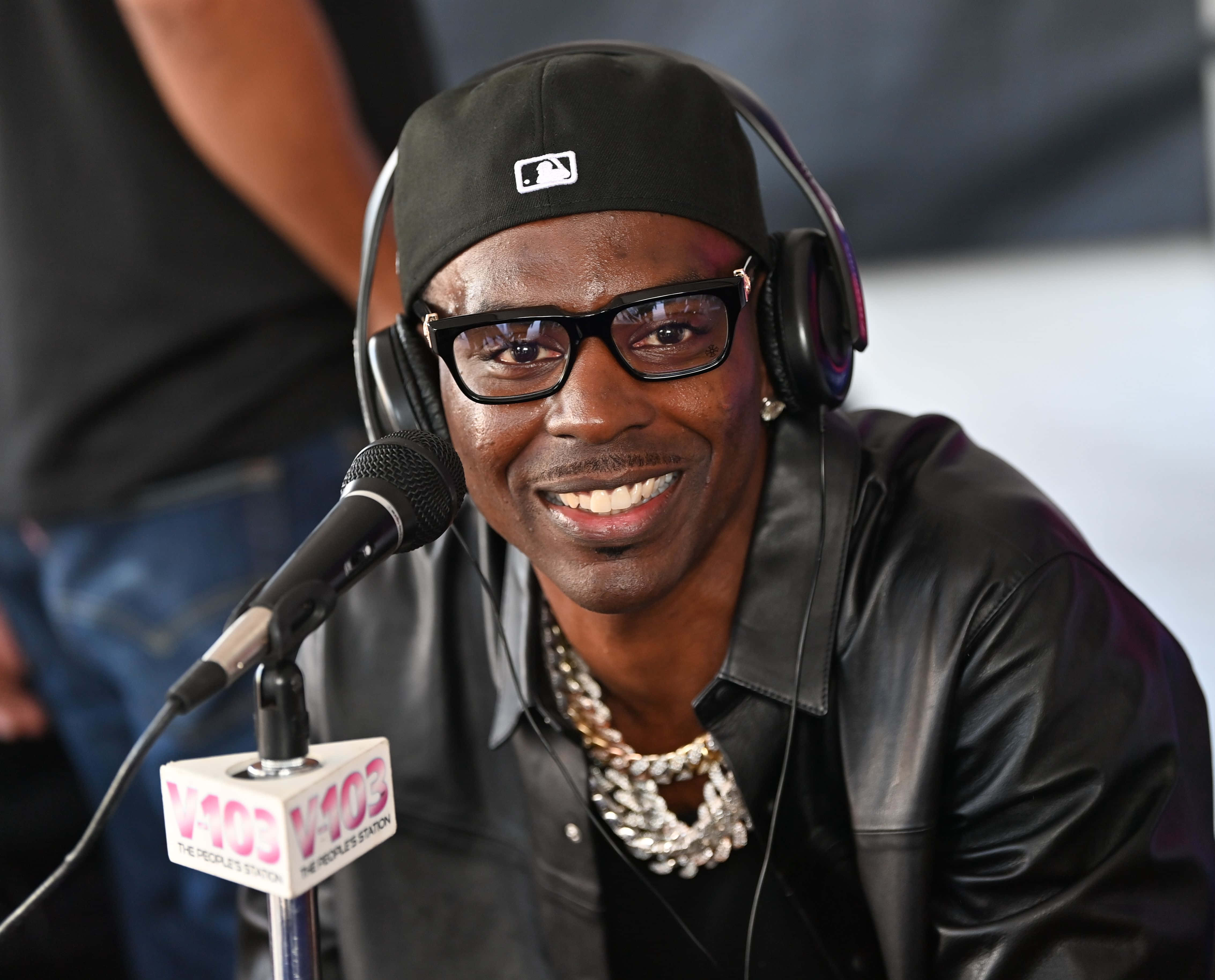 Young Dolph — 擊敗說唱界 壁紙 — 一個微笑的男人戴著黑色帽子和皮夾克，戴著耳機，對著V103電台咪高峰說話。