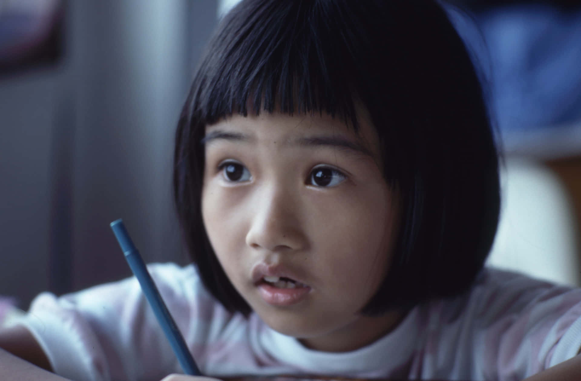  壁紙 — 一位留著深色波波頭和瀏海的年輕女孩，在柔和光線的室內環境中，拿著一支藍色筆，睜著大大的好奇眼睛看向鏡頭外。