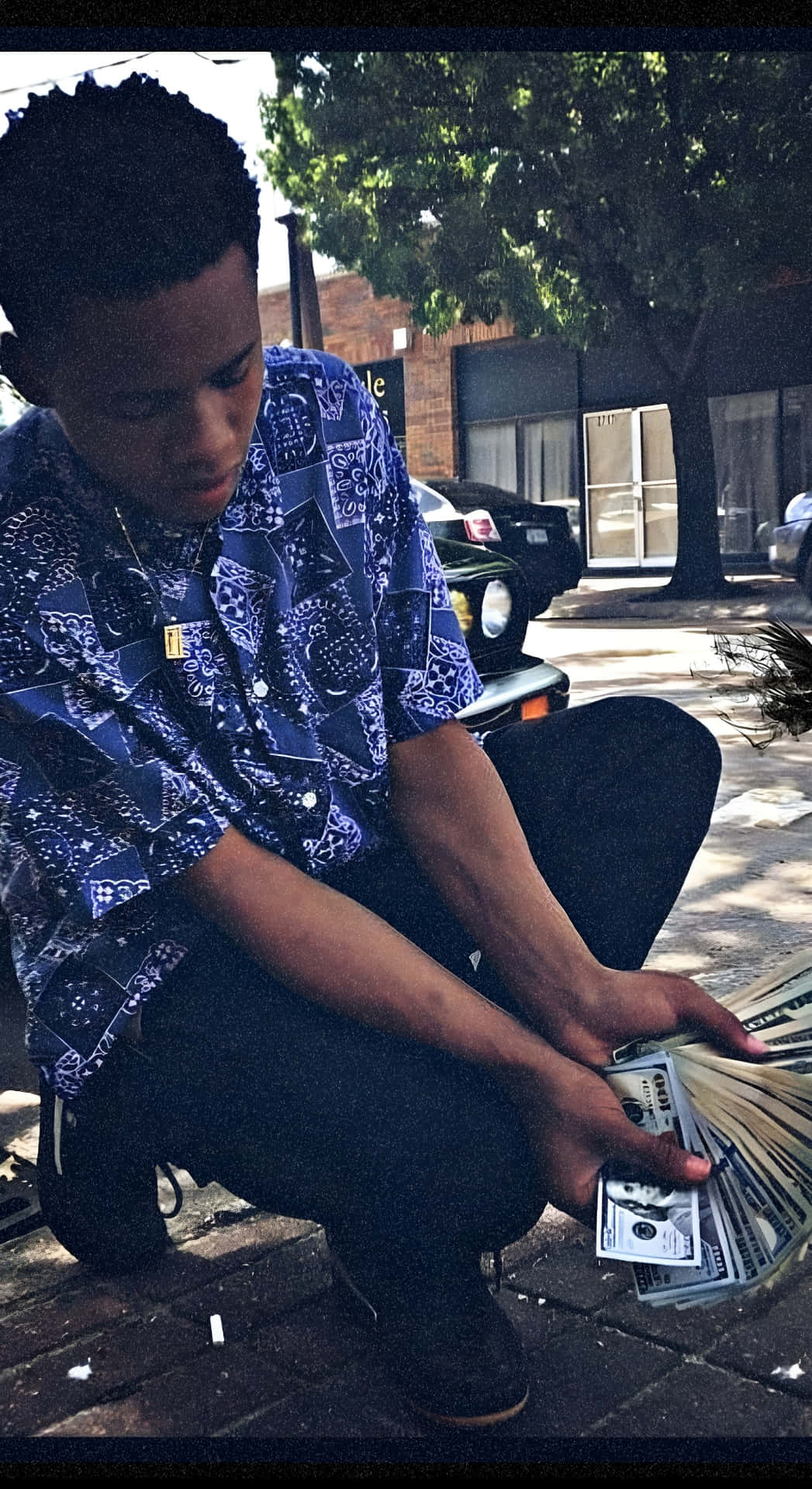  壁紙 — 一名身穿藍色圖案襯衫的年輕男子蹲在陽光普照的人行道上，專注地數著一疊現金。