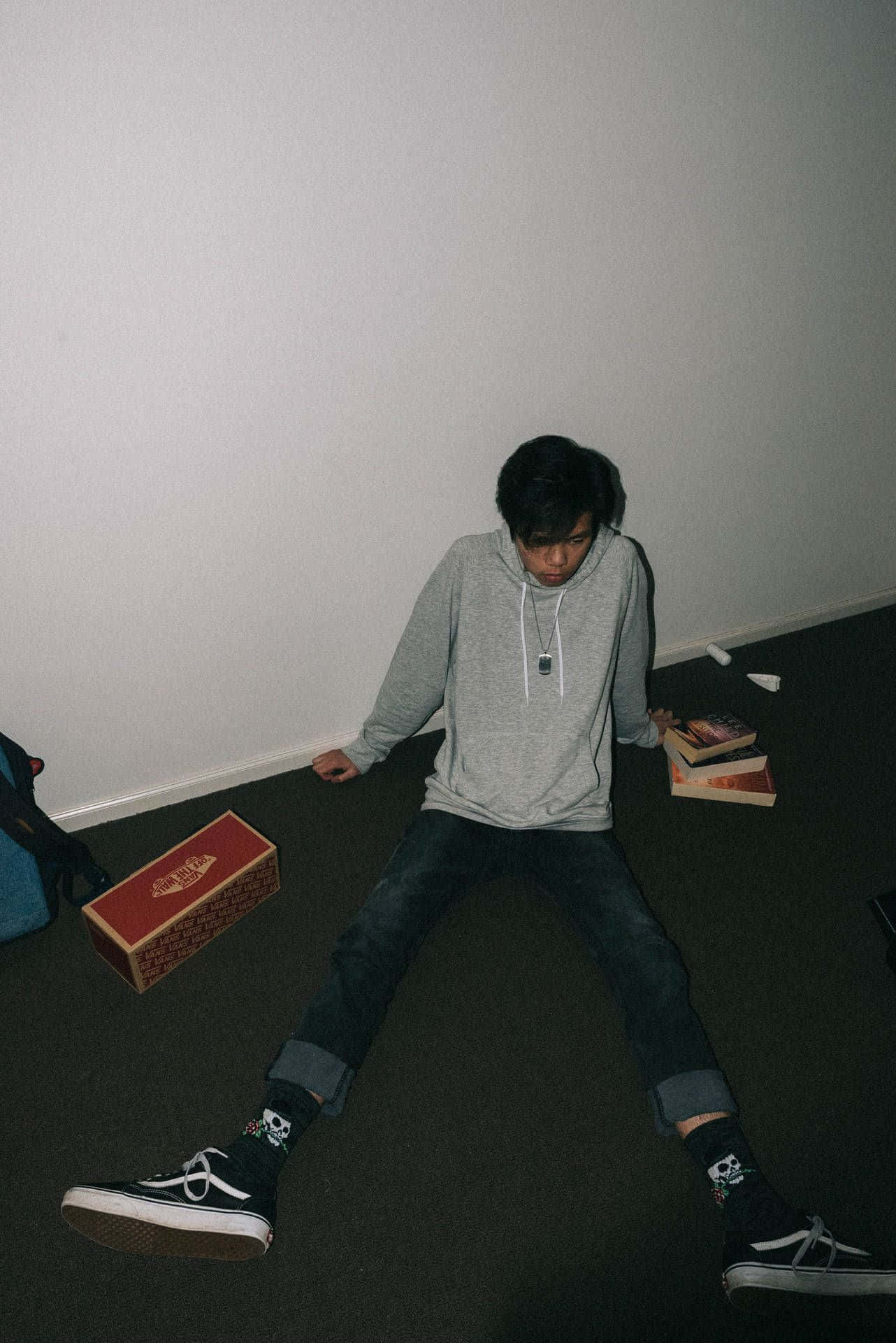  壁紙 — 一位身穿灰色連帽衫的年輕男子盤腿坐在深色地毯地板上，背靠純白牆壁，周圍散落著書籍和一個紅色盒子。