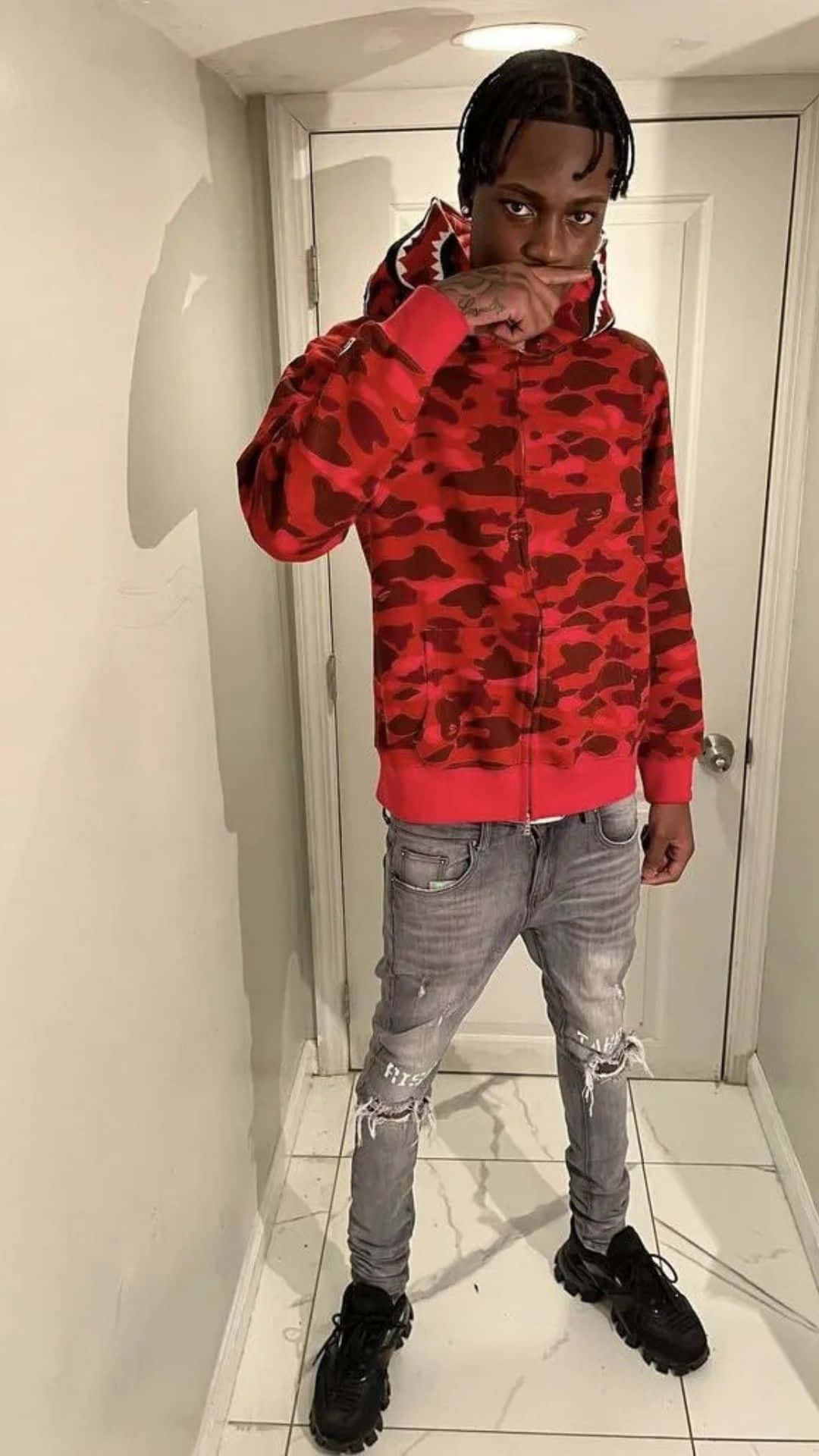 壁紙 — 一位身穿紅色迷彩連帽衛衣和破洞牛仔褲的年輕男子，在鋪有瓷磚、白牆和身後關著門的走廊裡自信地擺姿勢。