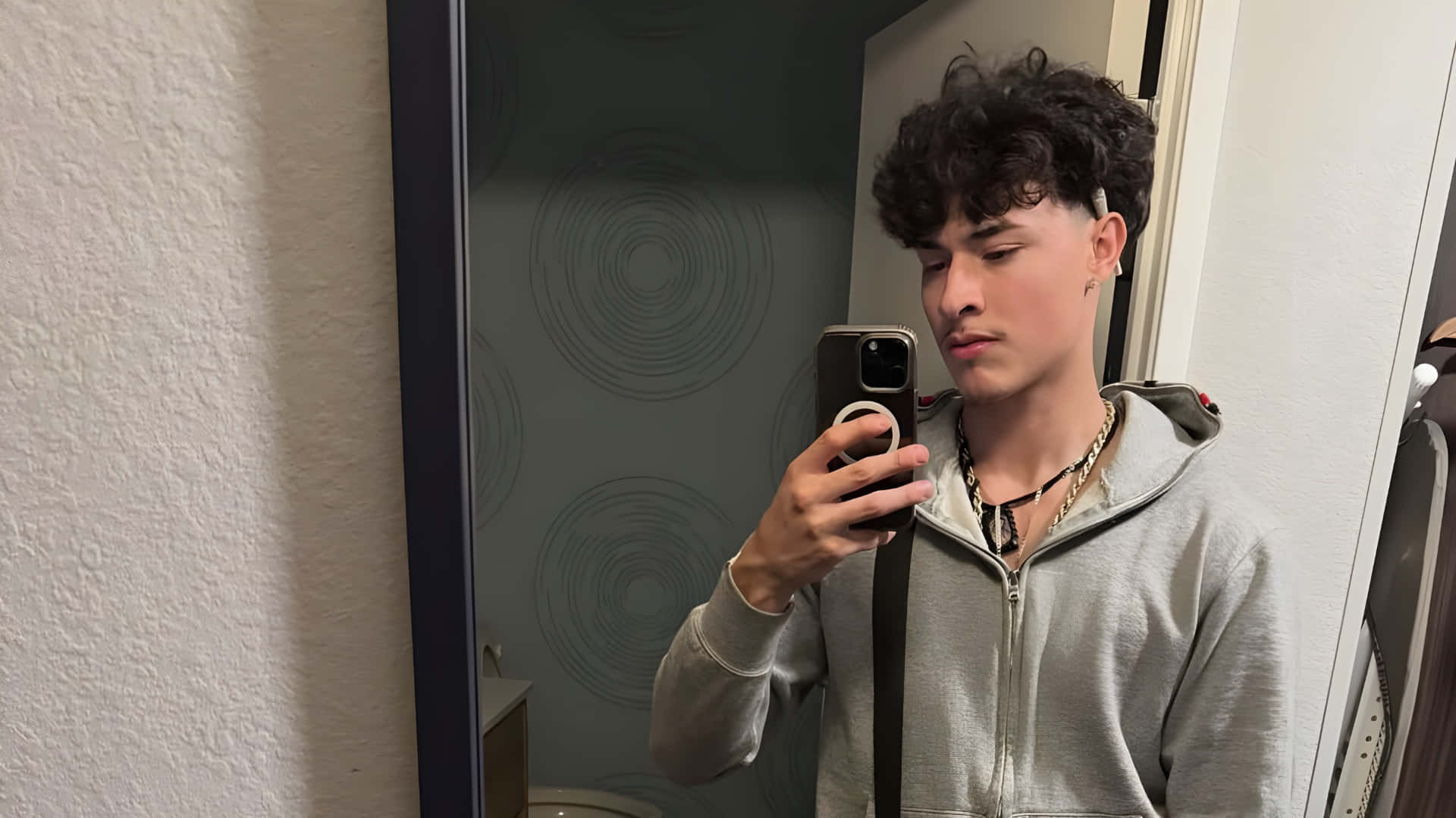  壁紙 — 一位捲髮年輕男子在浴室穿著灰色連帽衫和疊戴項鍊，對著鏡子自拍。