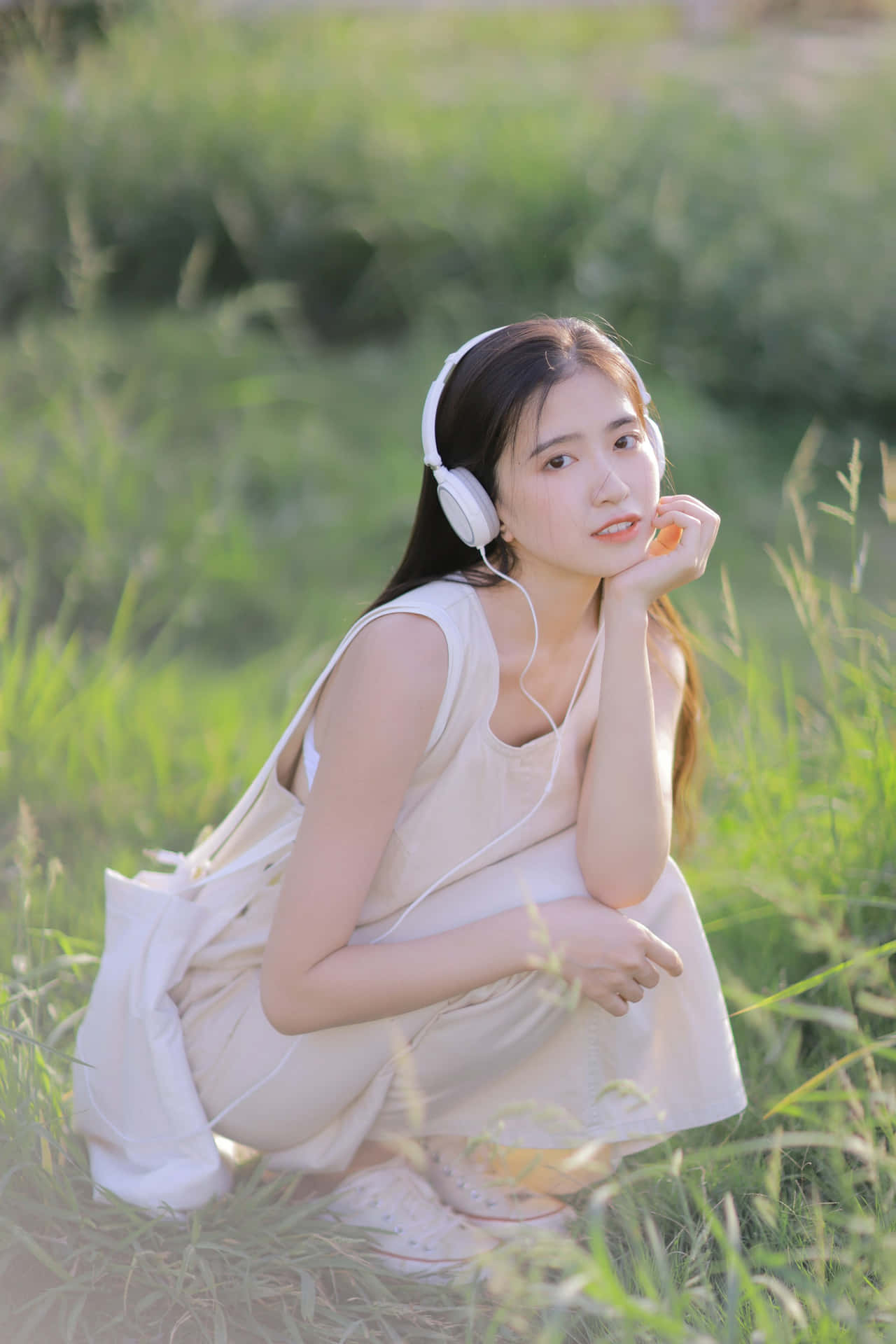  壁紙 — 一位身穿奶油色連衣裙的年輕女性坐在高高的草叢中，戴著白色耳機，對著鏡頭溫柔微笑。