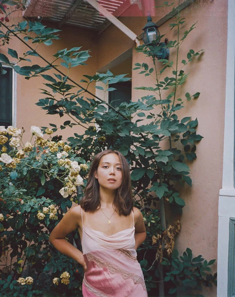  壁紙 — 一位身穿粉紅色連衣裙的年輕女子自信地站在茂密的綠葉和盛開的玫瑰中，背景是暖色調的建築牆壁。