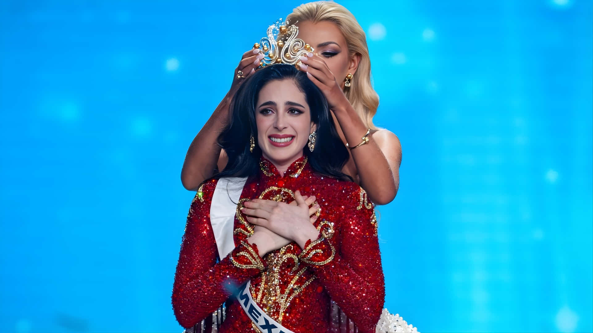  壁紙 — 一位年輕女士身穿閃亮紅色墨西哥選美禮服，由一位金髮女士在明亮藍色舞台背景下為她加冕。