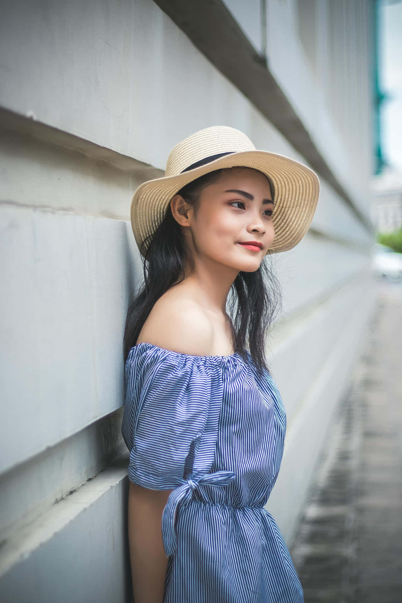  壁紙 — 一位戴草帽、身穿藍色條紋連衣裙的年輕女子，帶著溫柔的微笑靠在現代混凝土牆上。
