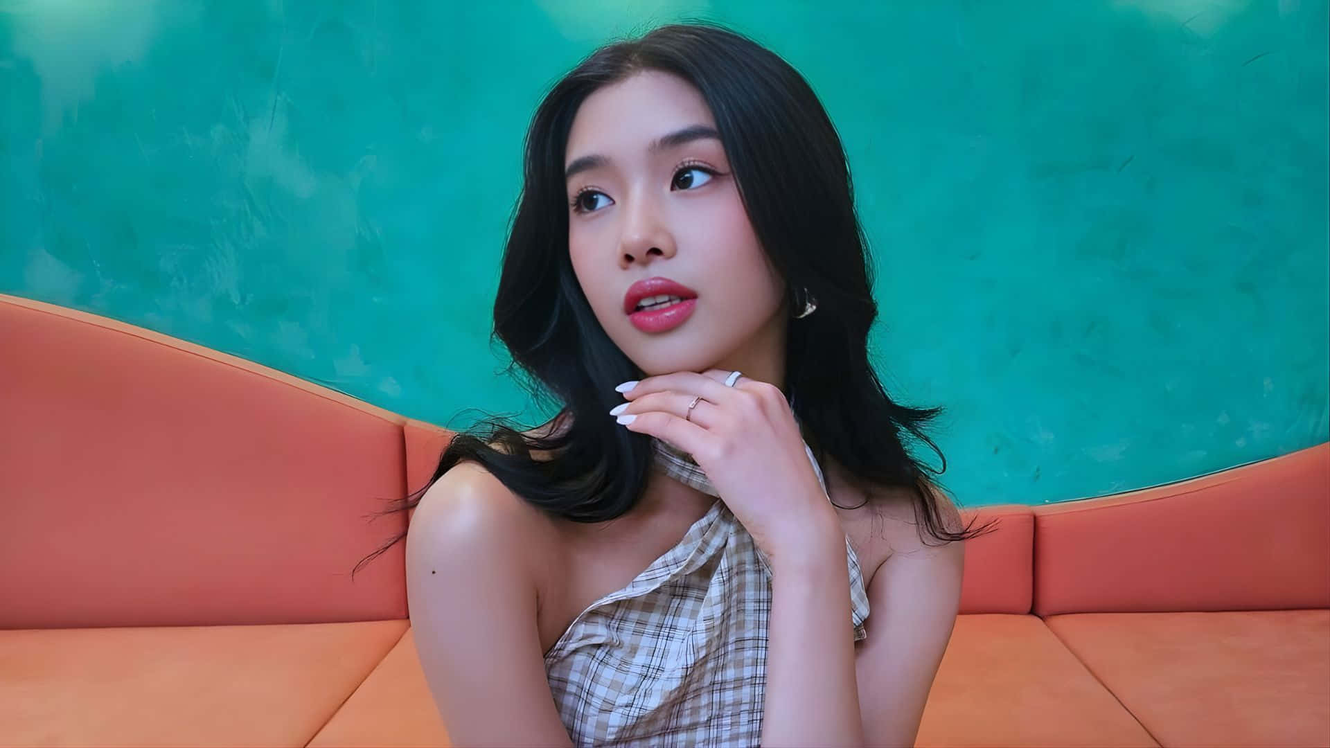  壁紙 — 一位深色頭髮的年輕女性坐在一張彎曲的橙色沙發上，背景是帶有紋理的青綠色牆壁，她抬頭凝視，表情若有所思。