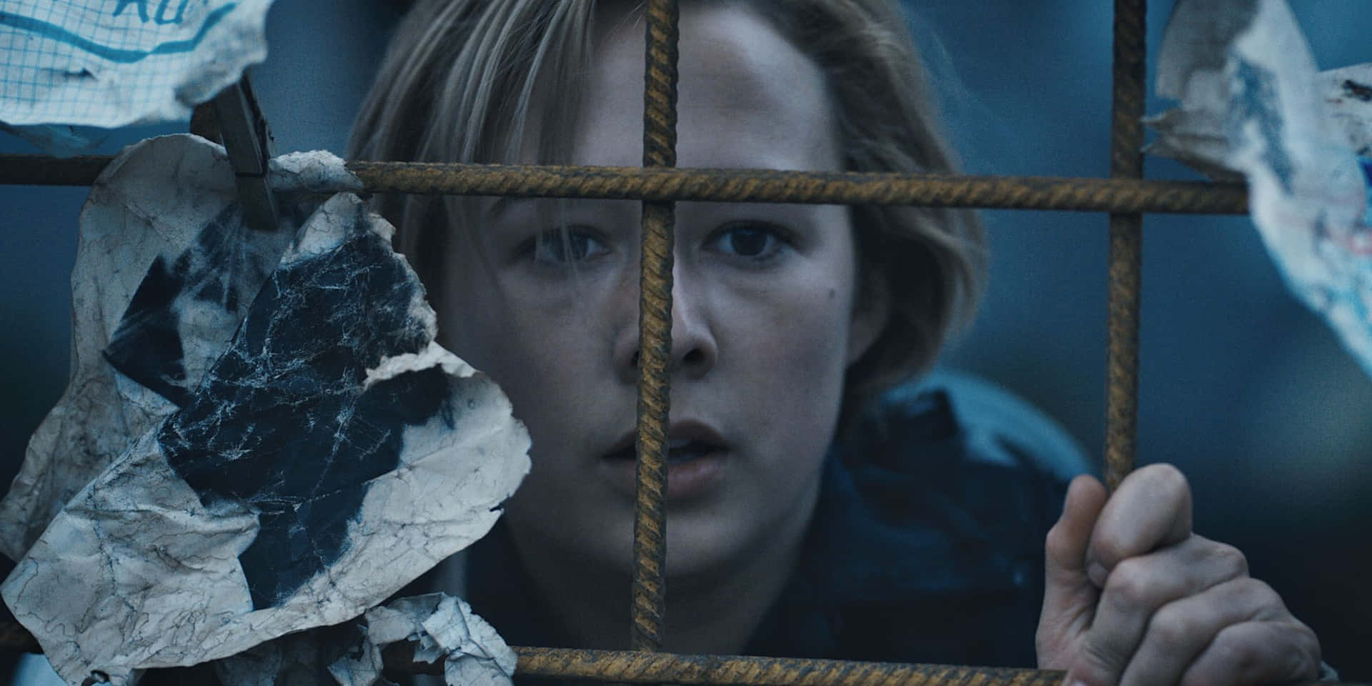  壁紙 — 一位年輕女子睜大眼睛透過生鏽的鐵欄杆凝視，她的手緊握金屬，撕碎的紙張在她周圍飄揚，場景昏暗藍光。
