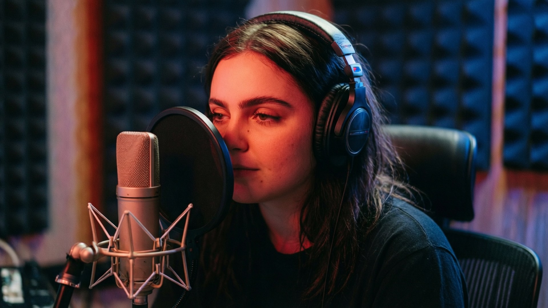 Studio Recording Session 壁紙 — 一位戴著耳機的年輕女性在溫暖的紅色燈光下，對著隔音泡沫牆前的錄音室麥克風唱歌。