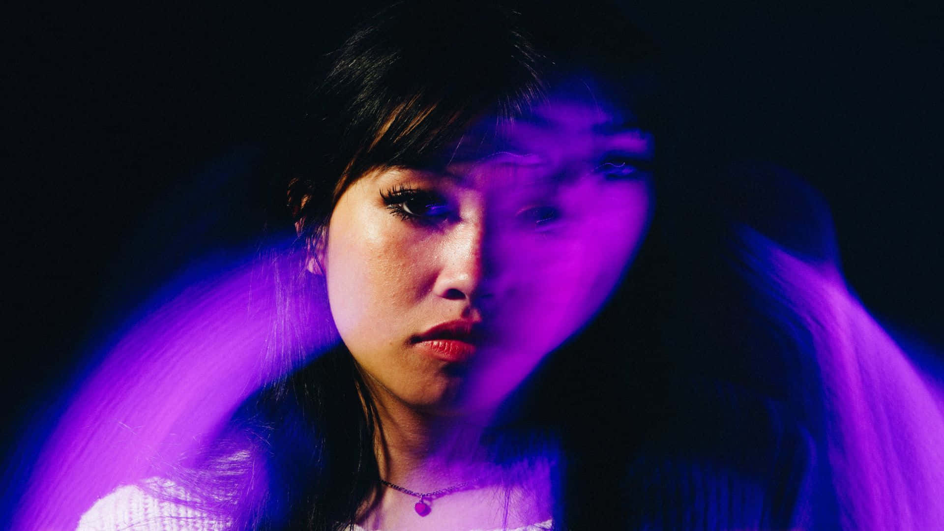  壁紙 — 一位黑髮年輕女子在黑色背景下，於鮮豔的紫色霓虹燈下凝視著。