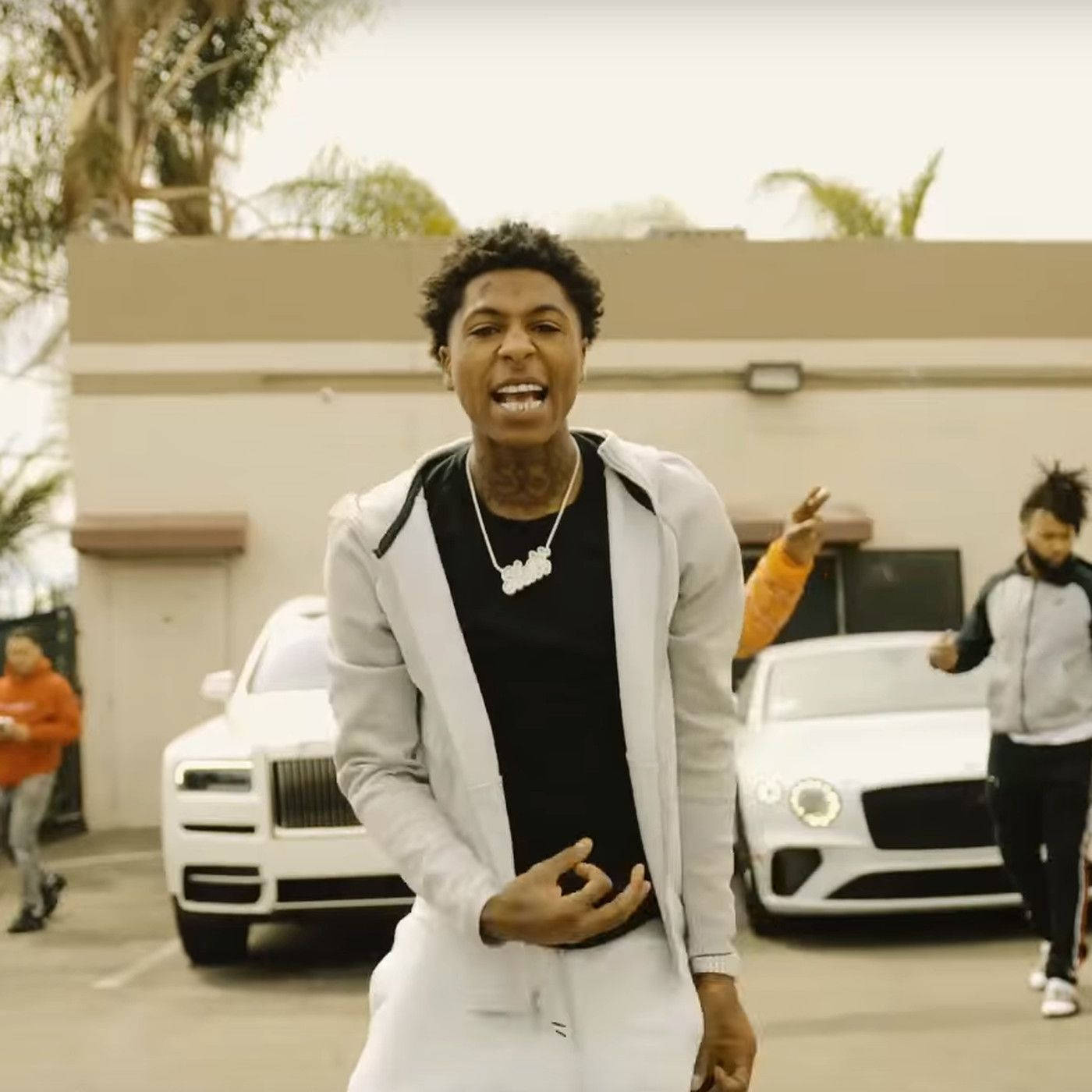Youngboy Never Broke Again追求成功 壁紙 — 一名身穿白色連帽衫和黑色襯衫的年輕男子在停車場兩輛豪華白色汽車前燦爛地微笑。