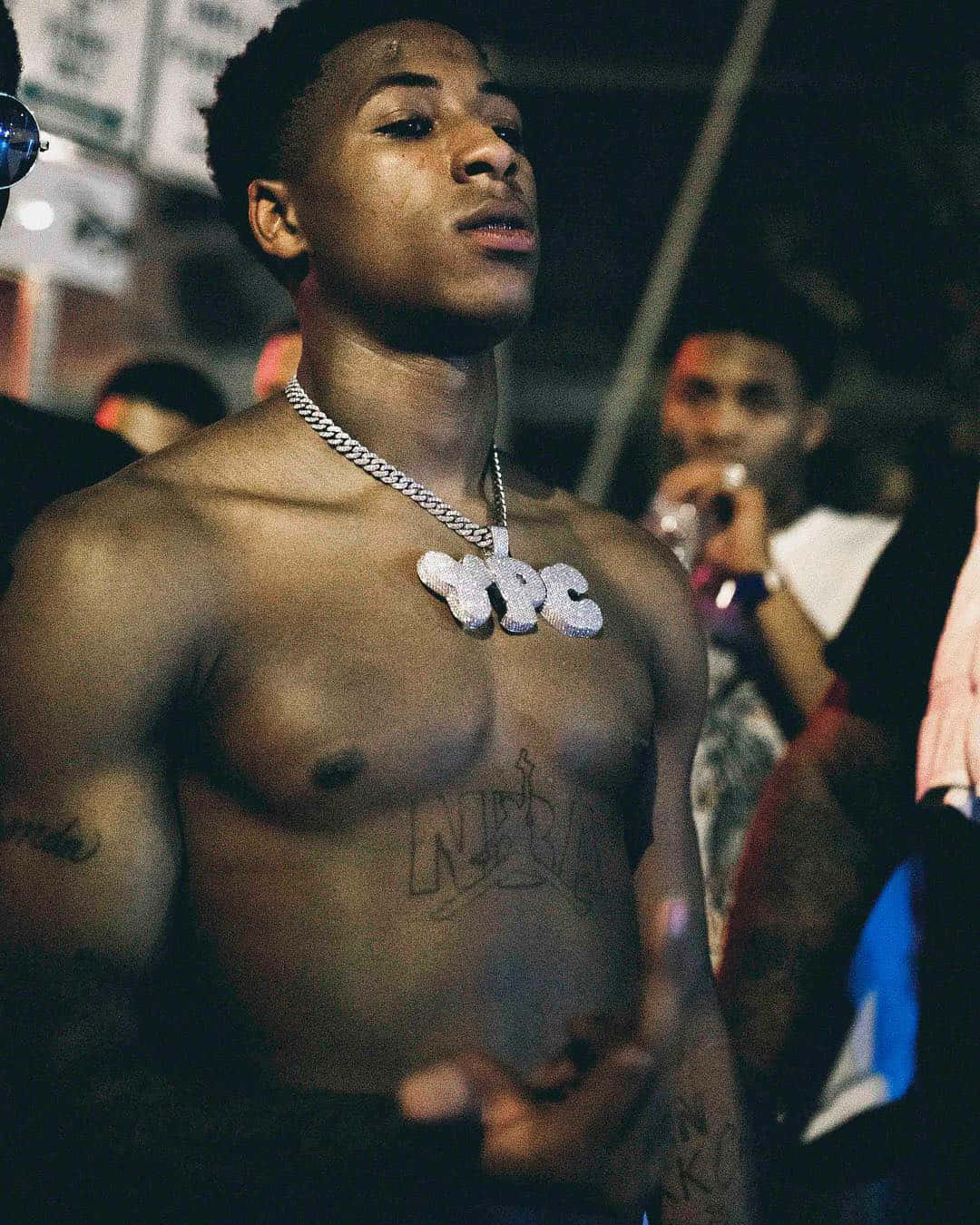 Youngboy Never Broke Again 壁紙 — 一個赤裸上身的年輕男子，戴著粗鏈和「NPC」吊墜，在昏暗的城市人群中向上看。