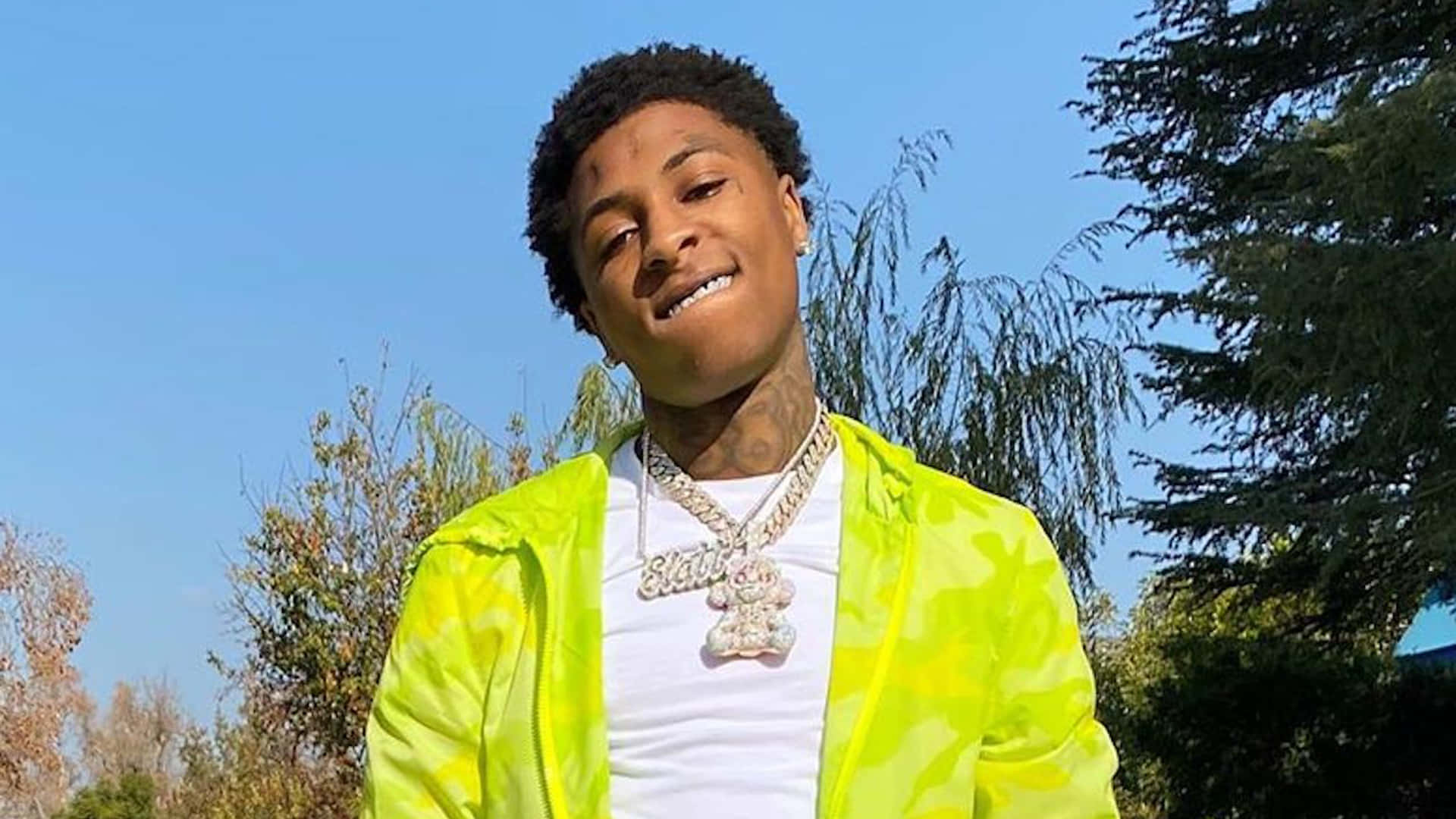 YoungBoy Never Broke Again 是饒舌歌手、歌手與詞曲創作人 壁紙 — 一位身穿亮眼螢光黃外套的年輕男子，在晴朗藍天和身後綠樹的襯托下自信地微笑。