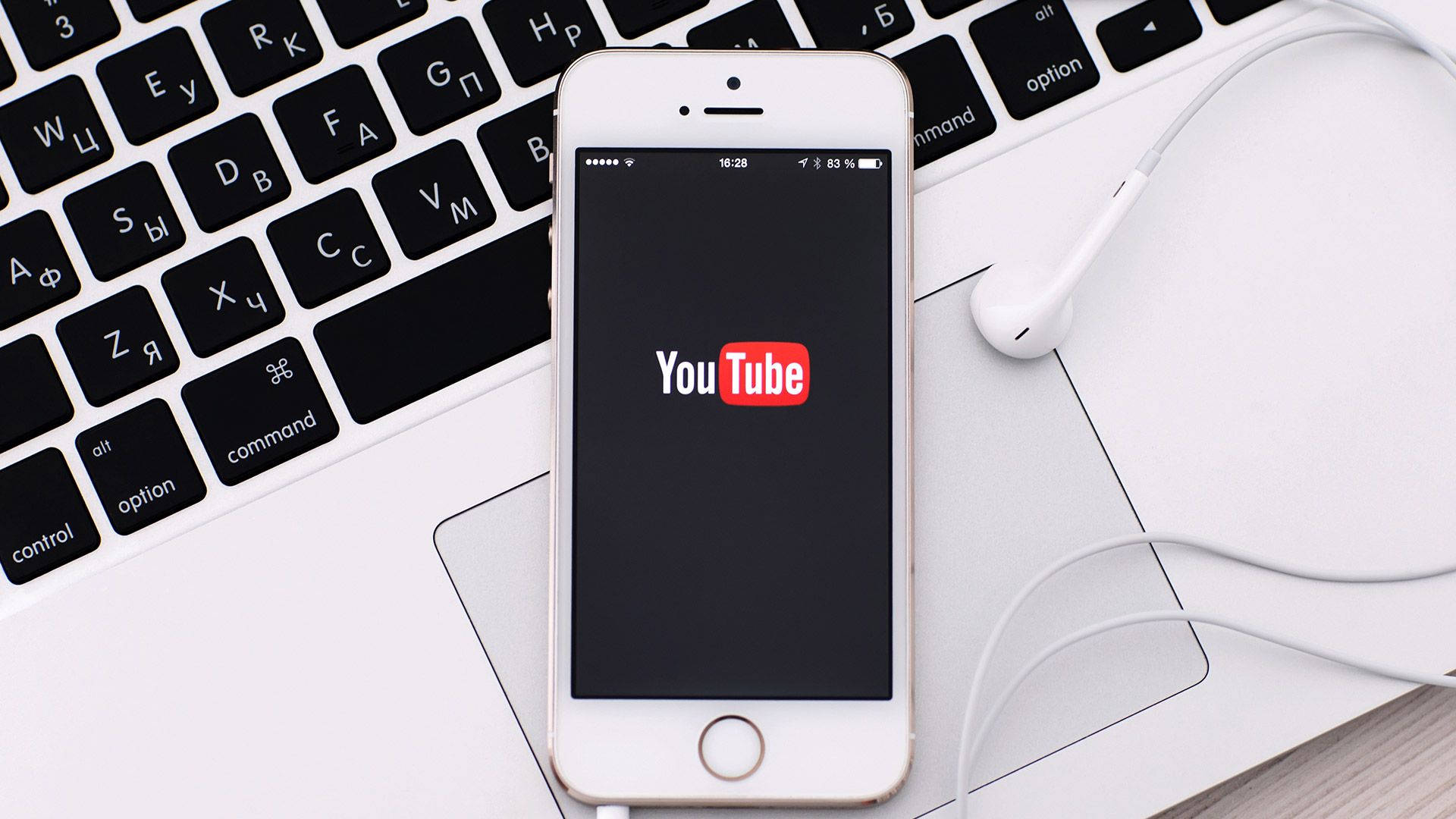 YouTube標誌顯示於手機螢幕上 壁紙 — 一部白色智能手機顯示YouTube標誌，放在手提電腦鍵盤上，旁邊有白色耳機。
