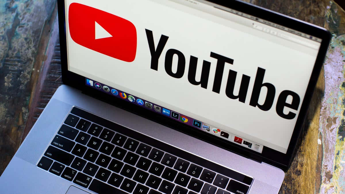 今天就在YouTube上讓你的內容被看見！ 壁紙 — 手提電腦屏幕上顯示 YouTube 標誌，背景為木桌。