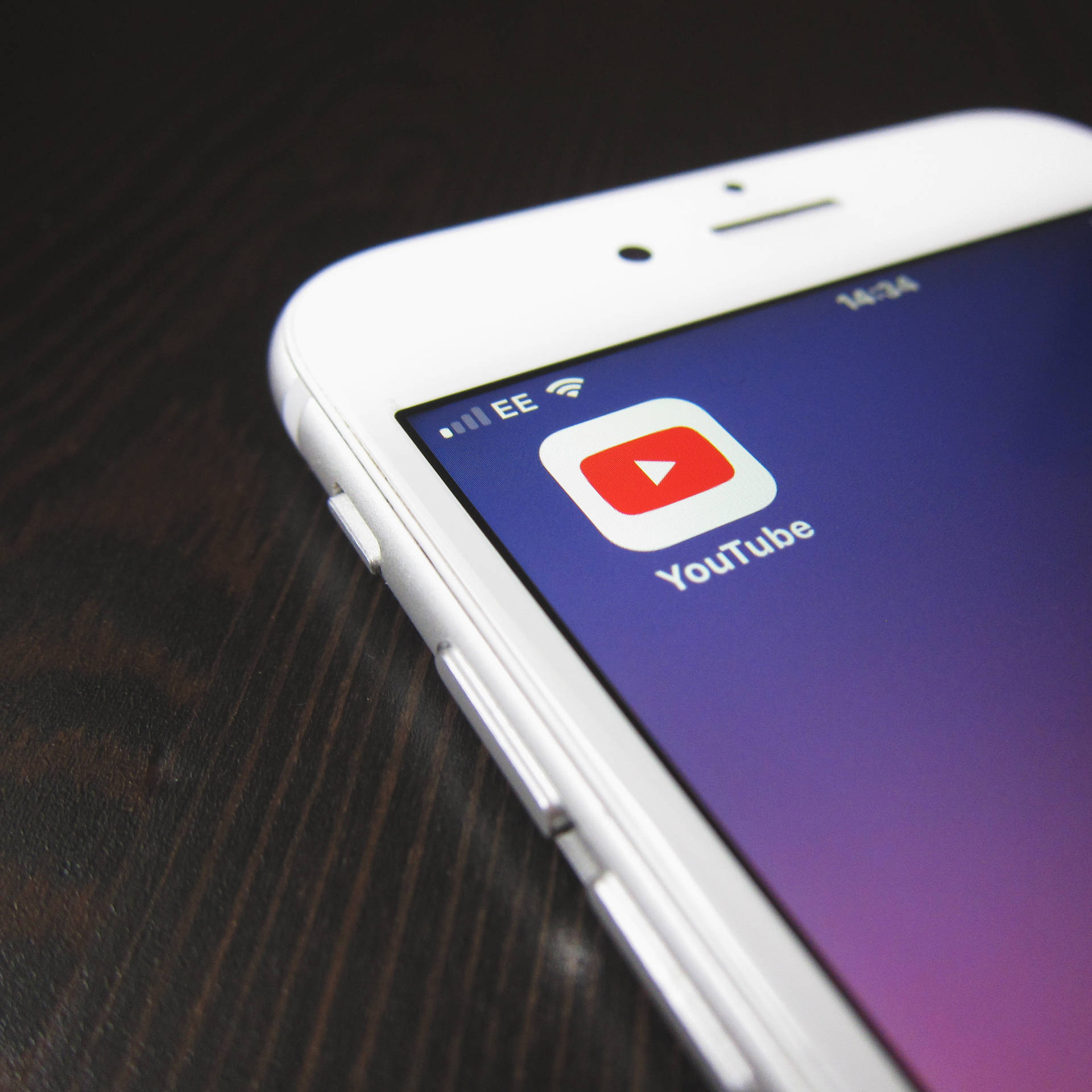 白色智能手機在紫色漸變背景上顯示 YouTube 應用程式，放在木桌上。