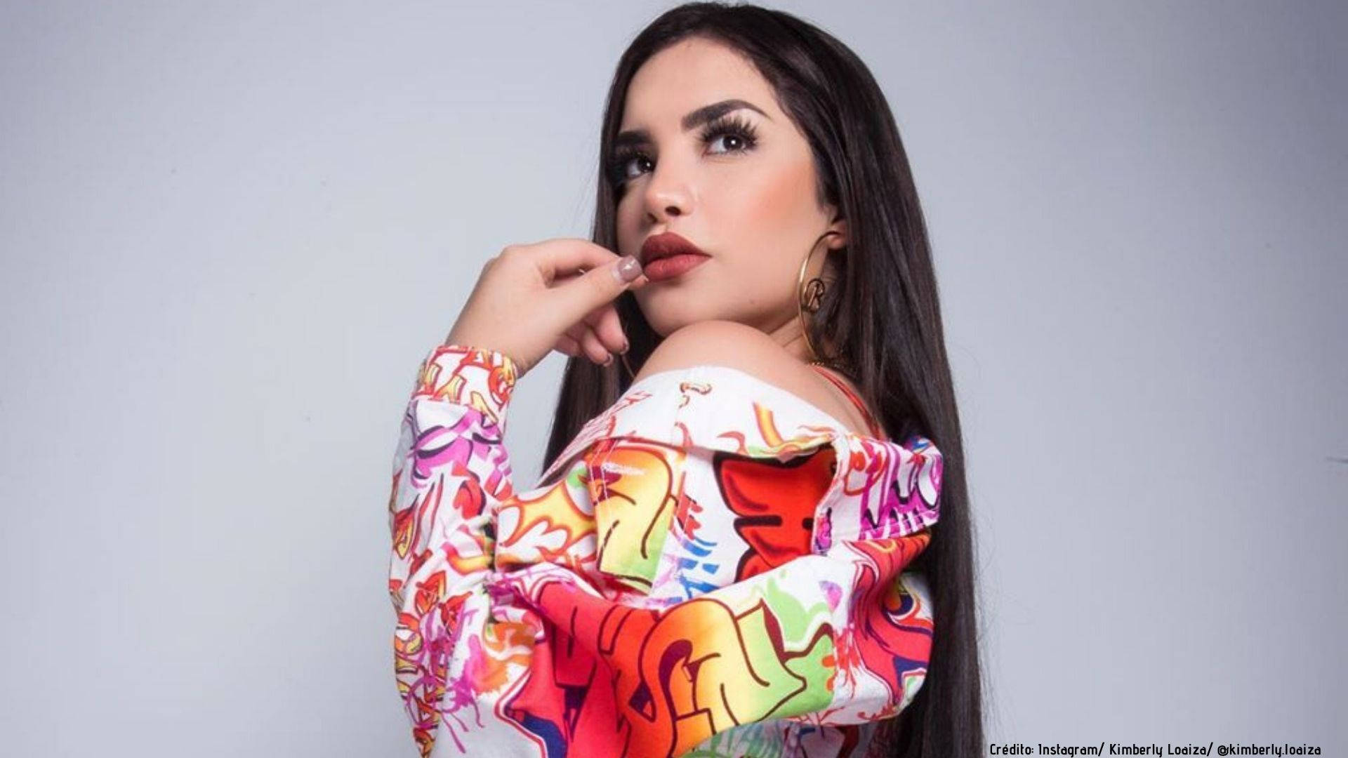 YouTube紅人Kimberly Loaiza身穿時尚夾克 壁紙 — 一位擁有長黑髮和大膽妝容的女性，在純灰色背景前，穿著充滿活力的塗鴉風格露肩上衣，自信地擺姿勢。