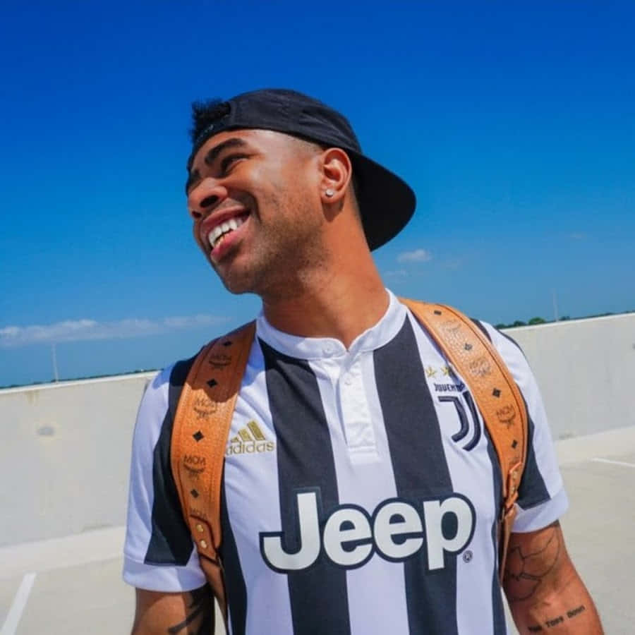 YouTuber Prettyboyfredo 背對藍天 壁紙 — 一個快樂的年輕男子穿著祖雲達斯足球球衣，在明亮的藍天下大笑，同時背著一個時尚的棕色背包。