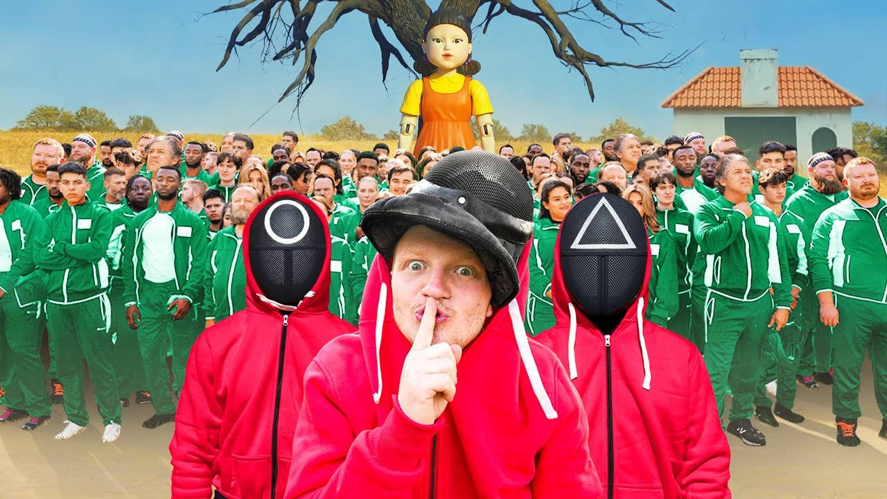 穿紅色連帽衫的男人將手指放在嘴唇上，站在穿綠色運動服的人群前，樹上有玩偶。